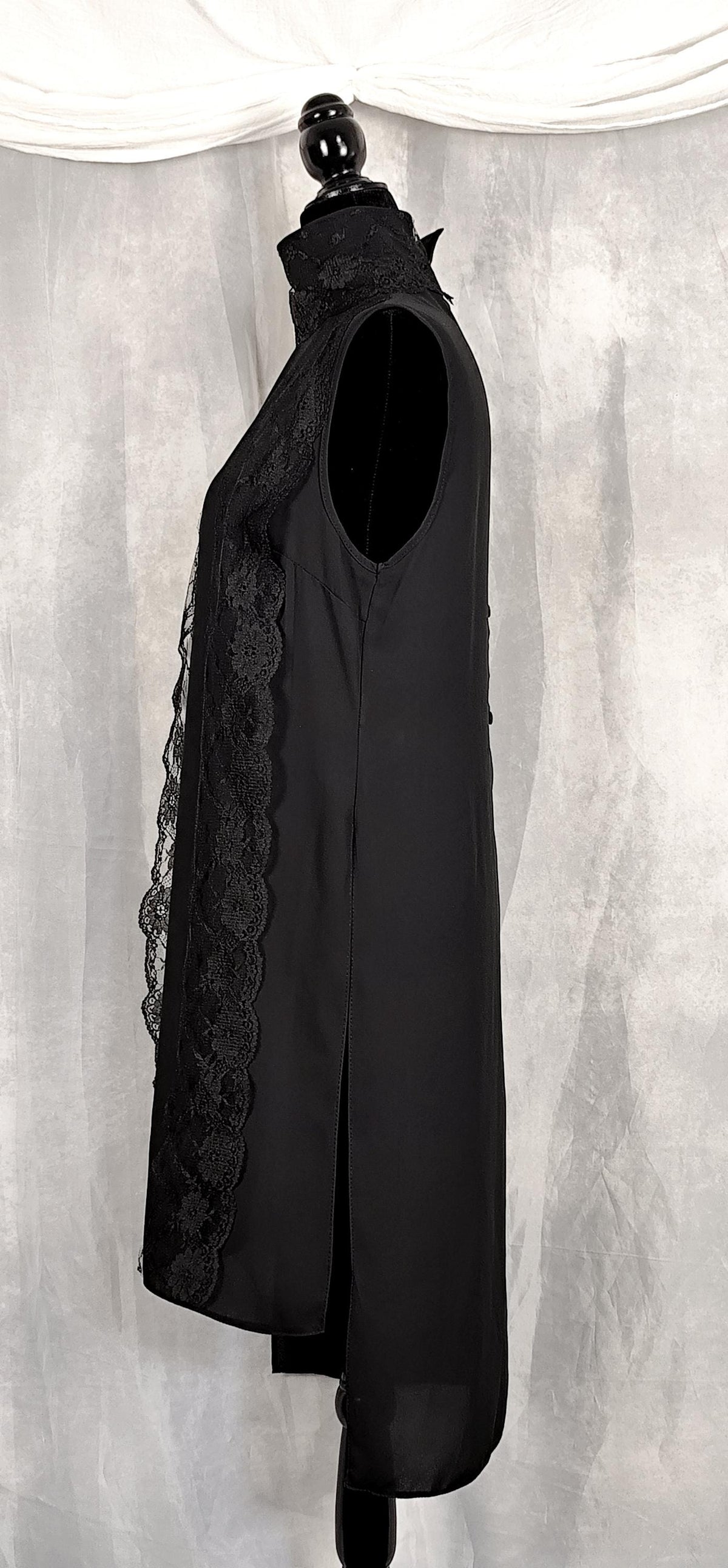 Side view of a sleeveless black long tunic with a high mock neck trimmed with lace and sheer lace side panels, displayed on a black mannequin against a gray draped fabric backdrop.