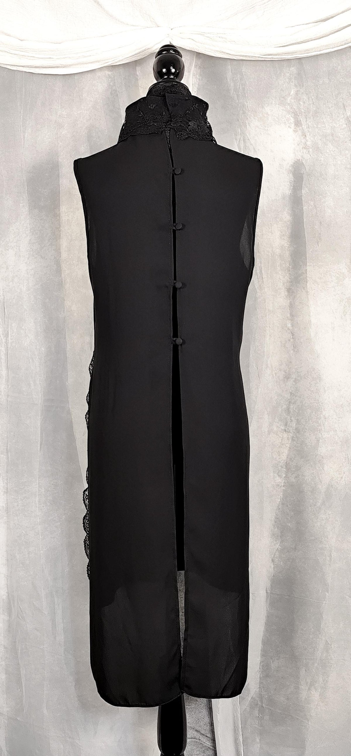 Back view of a sleeveless black long tunic with a high mock neck trimmed with lace and buttons on the back, displayed on a black mannequin against a gray draped fabric backdrop.