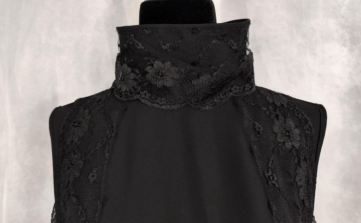 Close-up of a black sleeveless top with a high mock neck and floral lace detailing on the shoulders and collar, displayed on a black mannequin torso against a textured gray background.