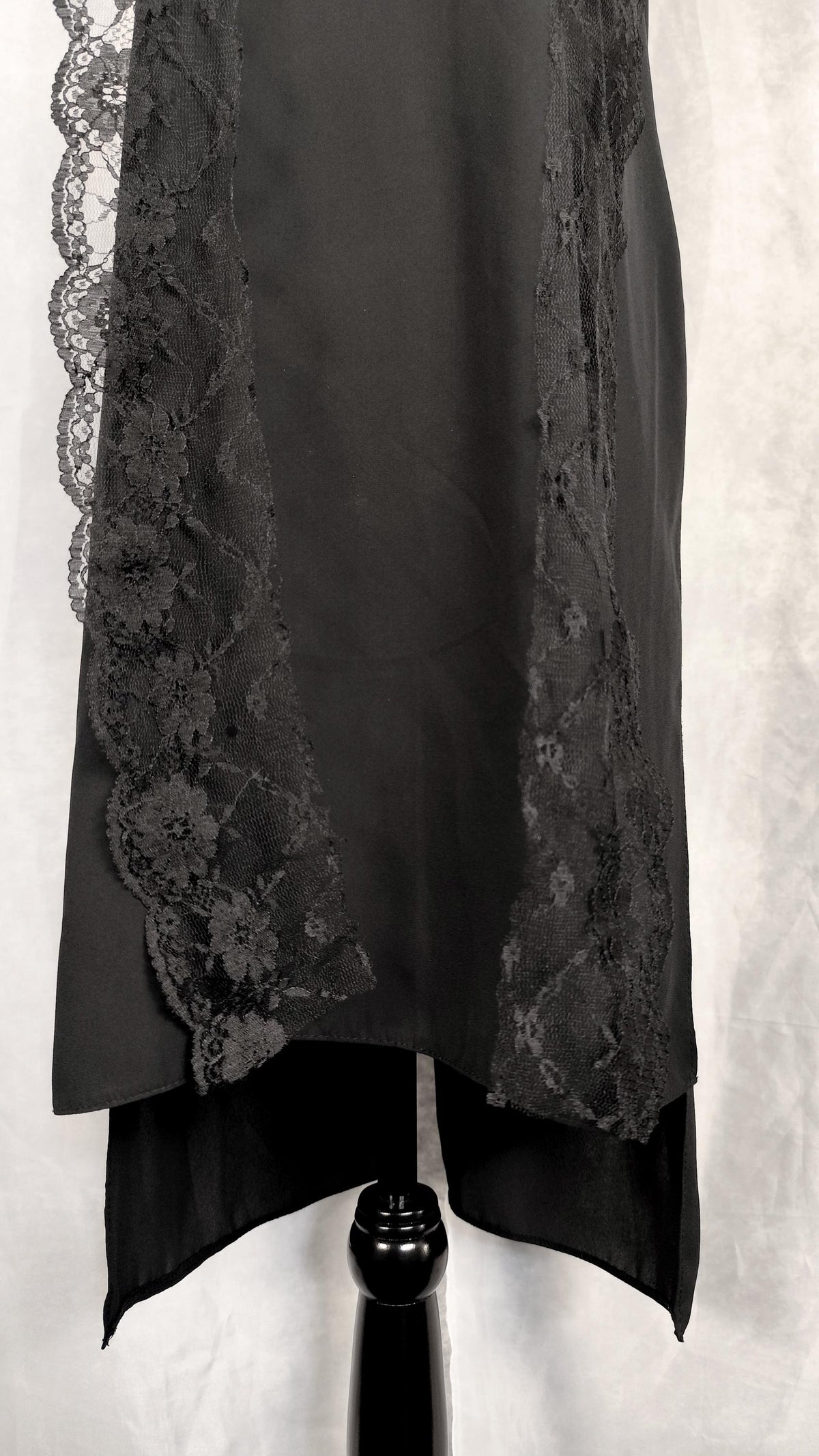 A black high-low tunic with intricate floral lace trim along the front edges, displayed on a black dress form against a textured light gray background.