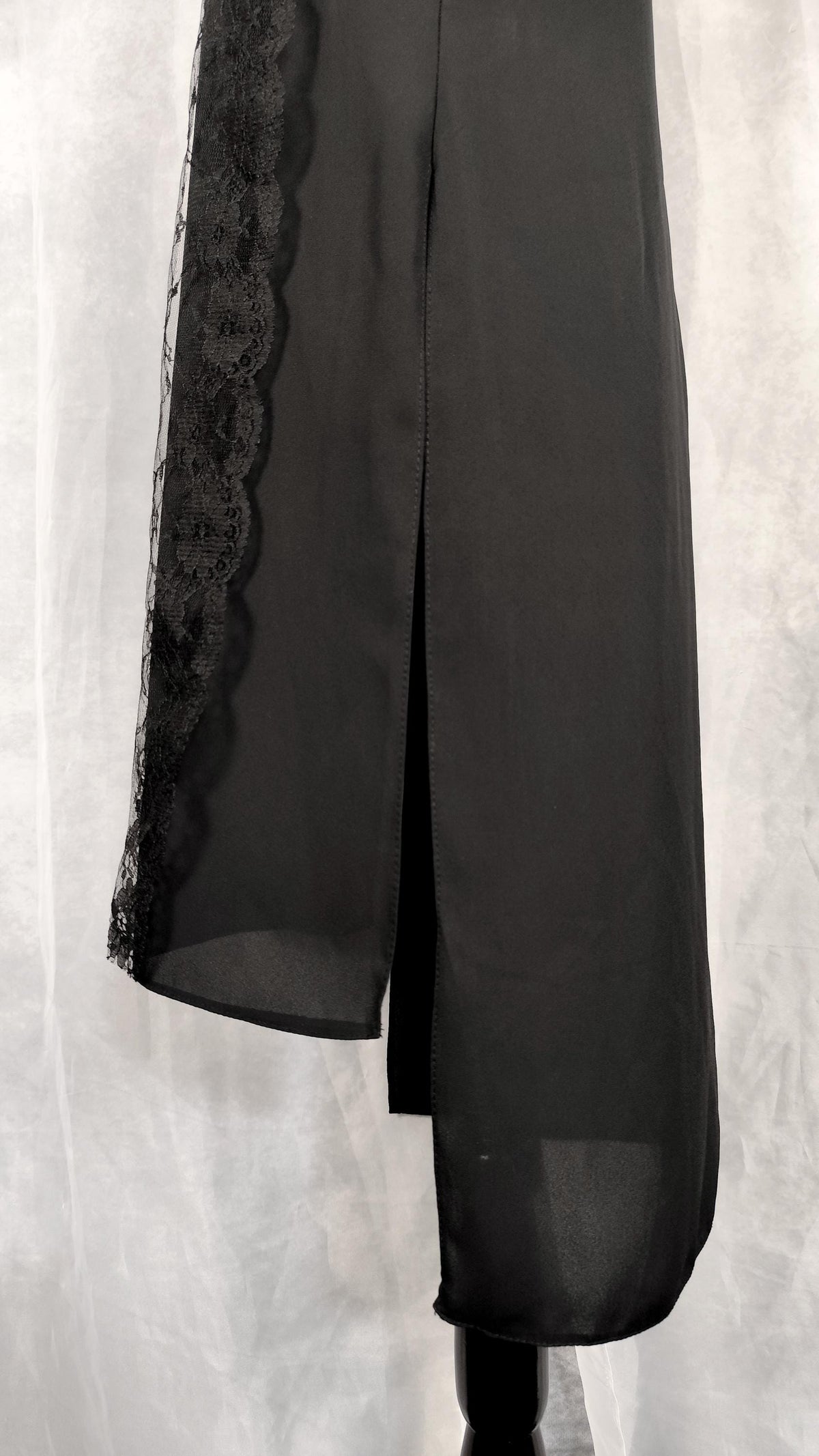 Close-up of a sleeveless black long tunic with a high mock neck trimmed with lace and sheer lace side panels, displayed on a black mannequin against a gray draped fabric backdrop.