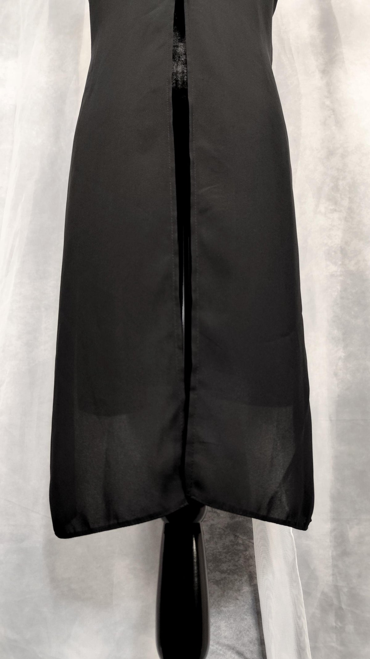 A black sleeveless tunic with a back slit, displayed on a black mannequin against a light gray draped fabric background. 