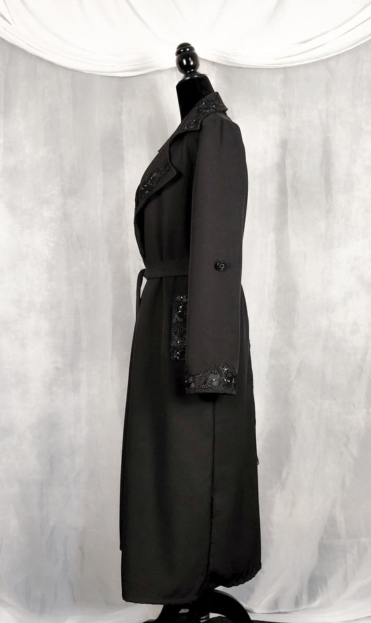 Side view of a black trench coat with decorative black lace trim on the collar, cuffs, and waist belt, displayed on a black mannequin against a draped white fabric backdrop.