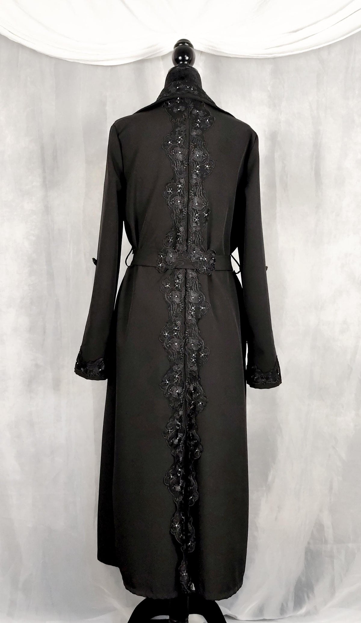 Back view of a black trench coat displayed on a mannequin against a draped gray fabric backdrop. The trench coat features a high collar, long sleeves, and a decorative vertical embroidered or lace panel running down the center front.