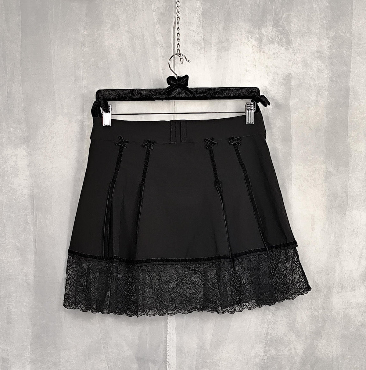 Back view of a black pleated mini skirt with lace trim and tie details, hanging on a black hanger against a textured gray wall.