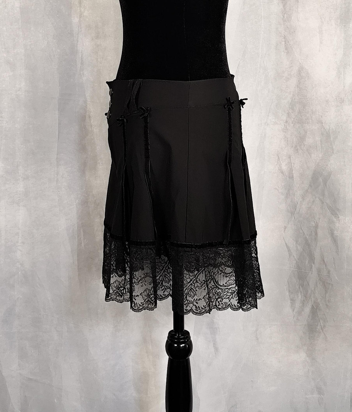 Side view of a black pleated skirt with a lace hem and side bows, displayed on a black dress form against a textured gray backdrop.