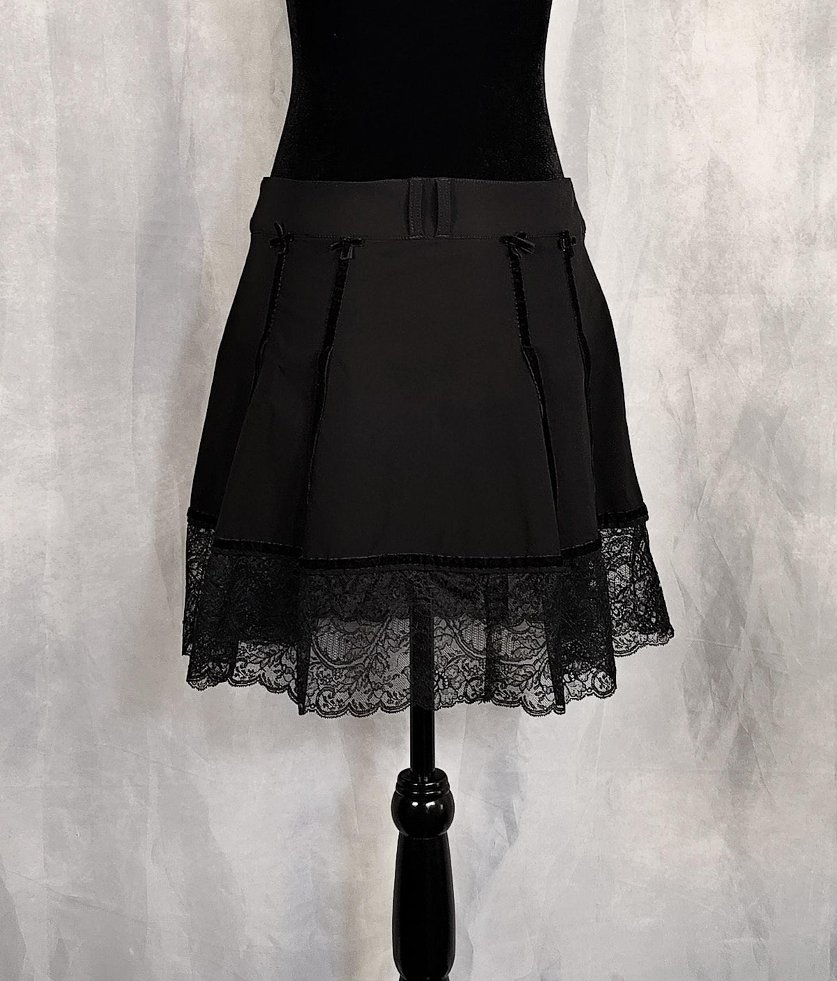 Back view of a black pleated skirt with a lace hem, displayed on a black dress form against a textured gray backdrop.
