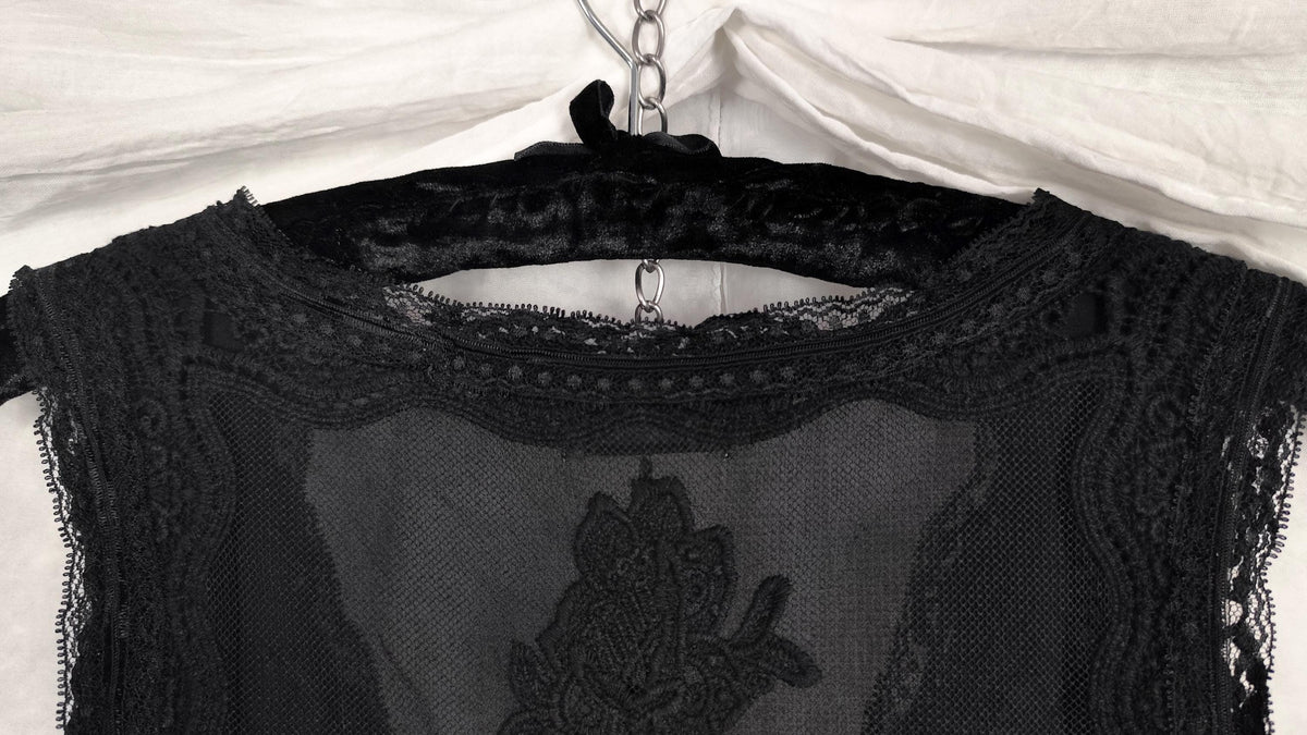 A black sheer lace midi dress with a floral applique detail, hanging on a black velvet hanger against a white fabric backdrop.