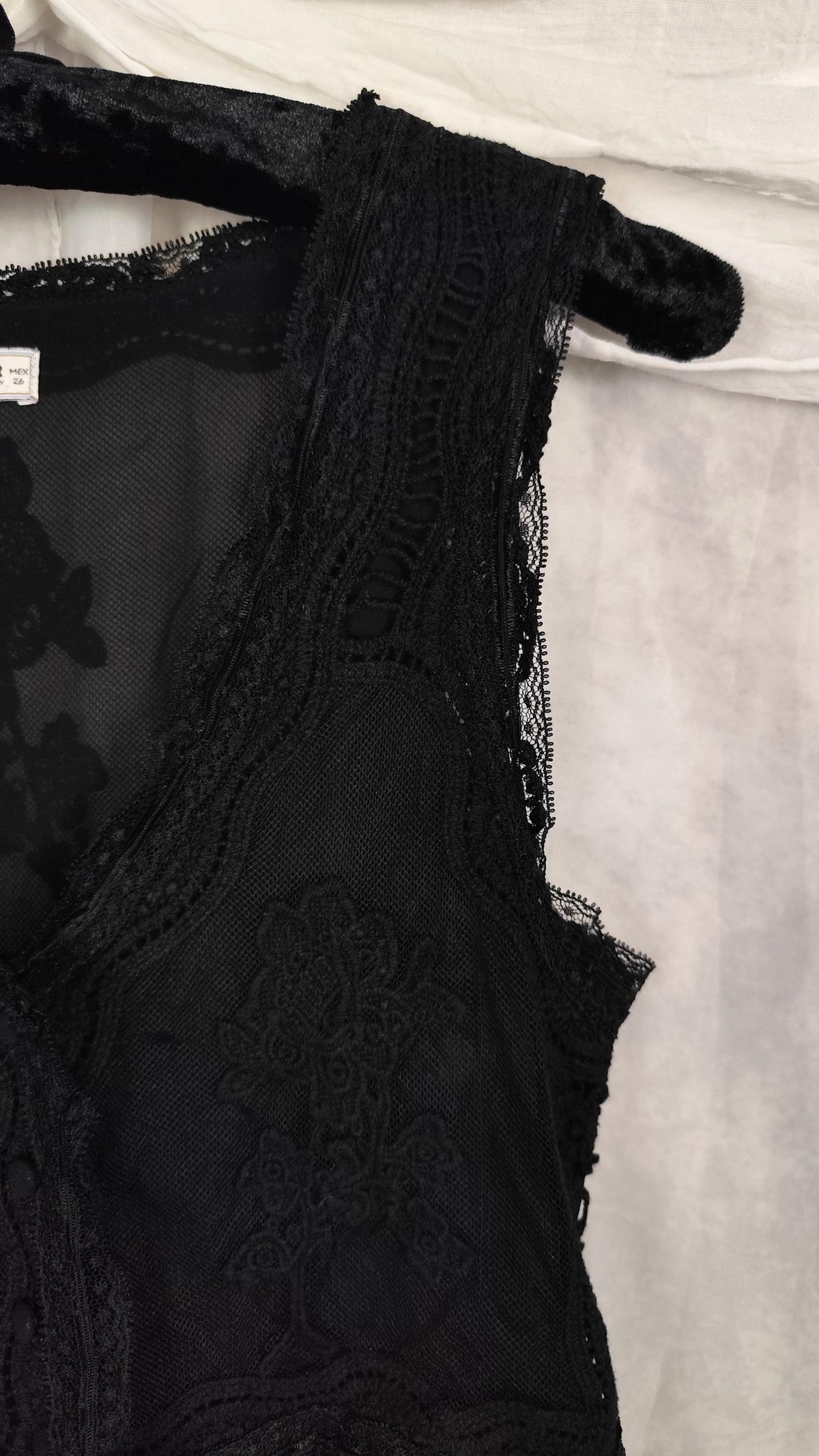 Close-up of a black lace sleeveless midi dress with floral embroidery and sheer mesh, hanging against a light fabric backdrop.