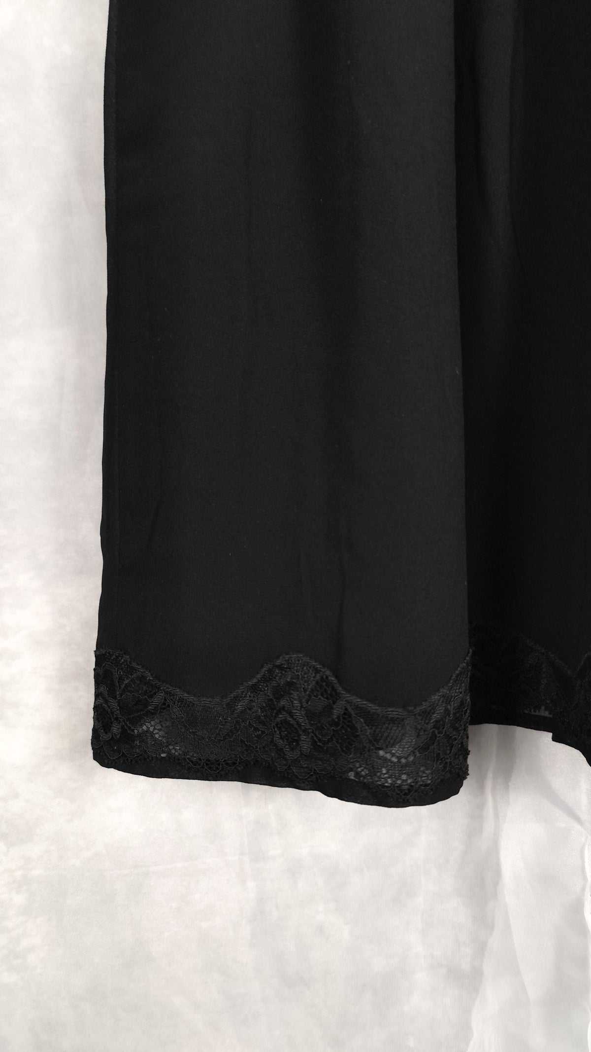 A close-up of a black midi dress featuring a delicate black lace trim along the hem, displayed against a white fabric backdrop.