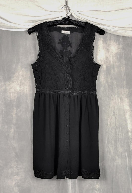 Front view of a sleeveless black dress with lace detailing on the bodice and a button-front overlay, hanging on a black hanger against a gray textured backdrop with draped white fabric at the top.