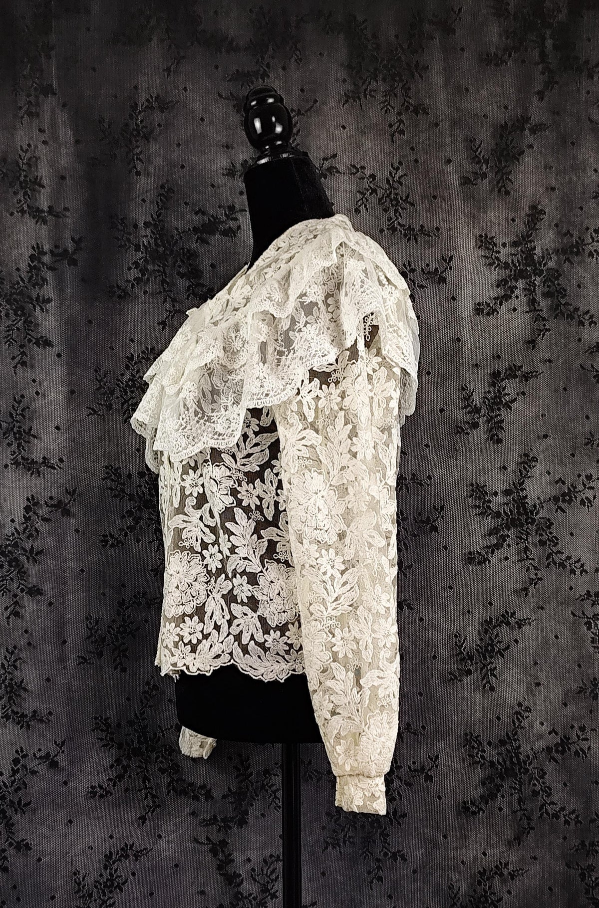 Side view of a off-white lace ruffled blouse with long sleeves, displayed on a black mannequin torso against a dark textured floral backdrop.