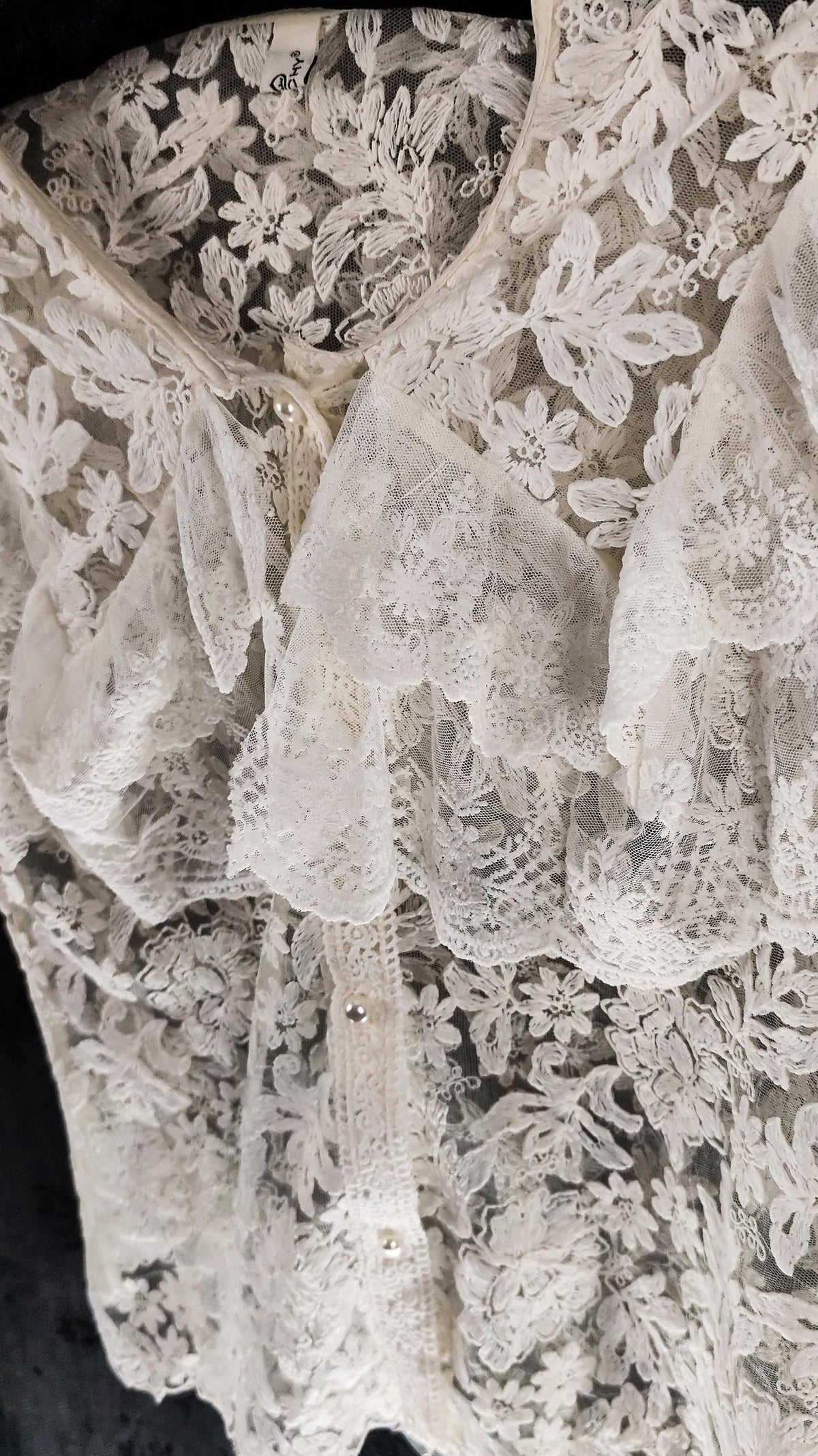 Close-up of a off-white floral lace blouse with ruffled trim and a pearl-shaped button front, shown against a dark background.