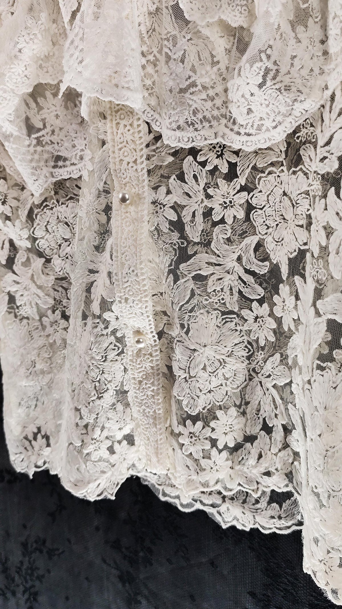 Close-up of a off-white floral lace blouse with ruffled collar and small pearl-shaped buttons, laid against a dark textured background.