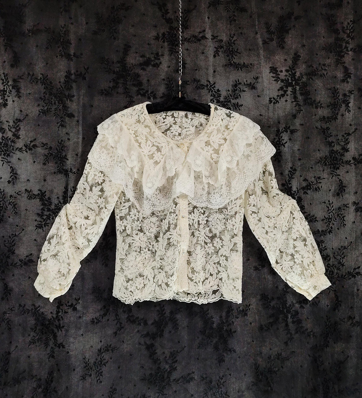 Front view of an off-white lace blouse with a large ruffled collar and long sleeves, hanging on a black hanger against a dark, textured background.