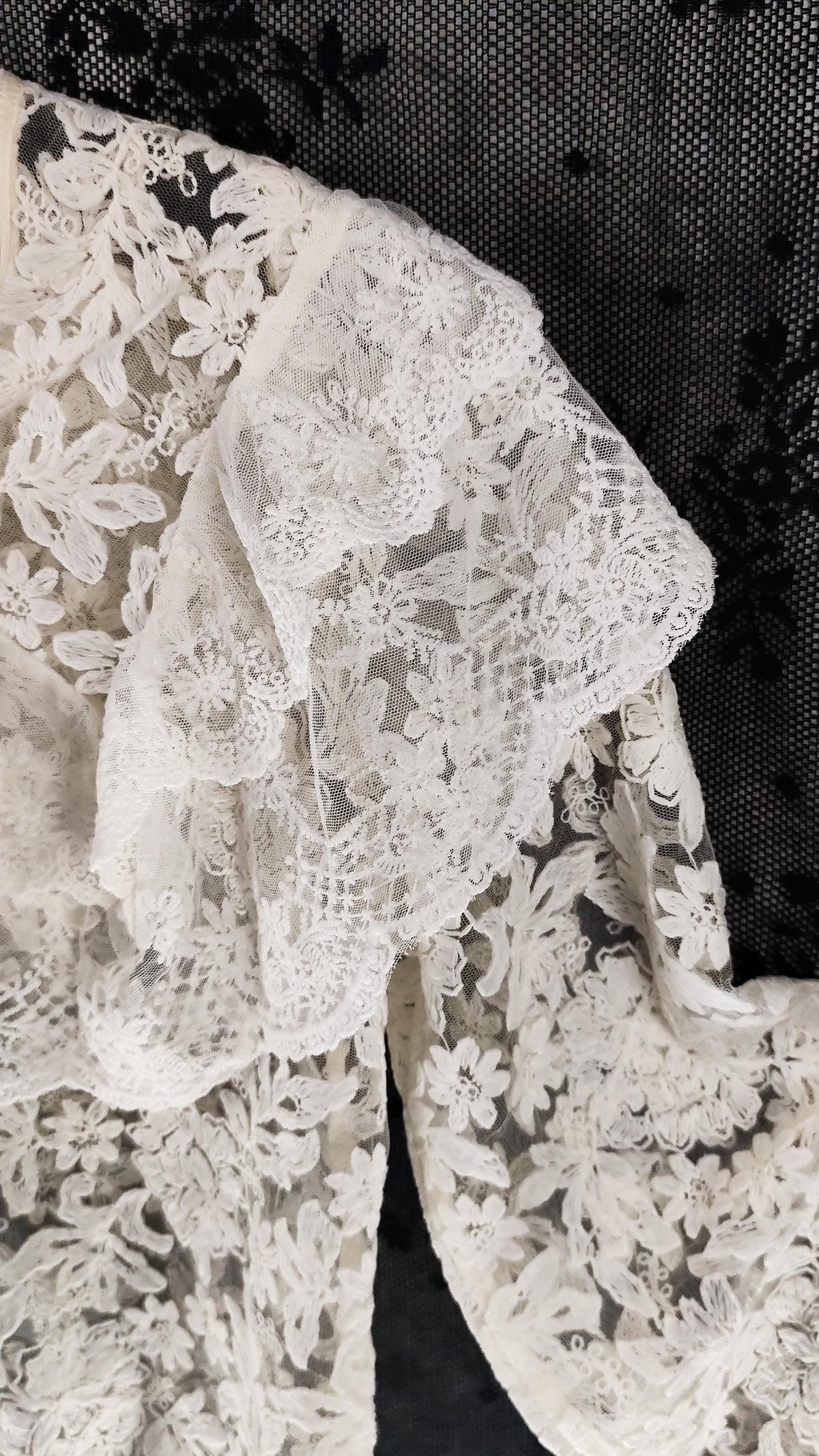 Close-up of a off-white floral lace blouse draped over a black mesh background, showing the detailed lace pattern and texture.
