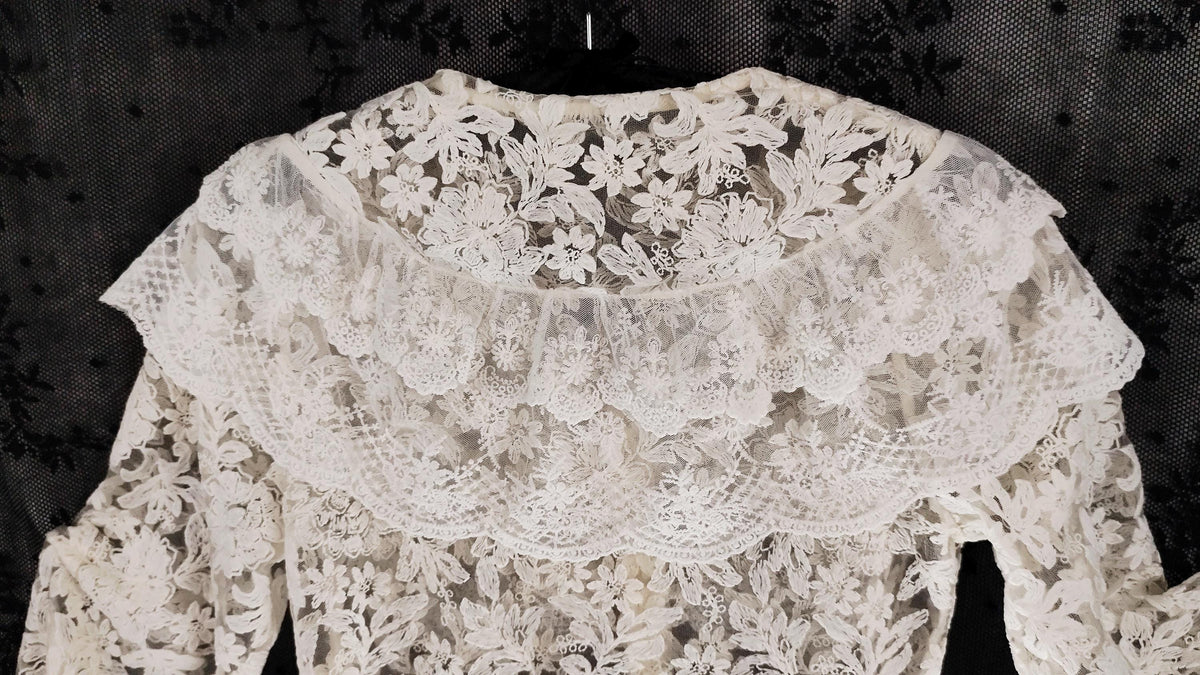 An off-white lace blouse with intricate floral embroidery and a wide ruffled collar, displayed against a dark patterned fabric background.