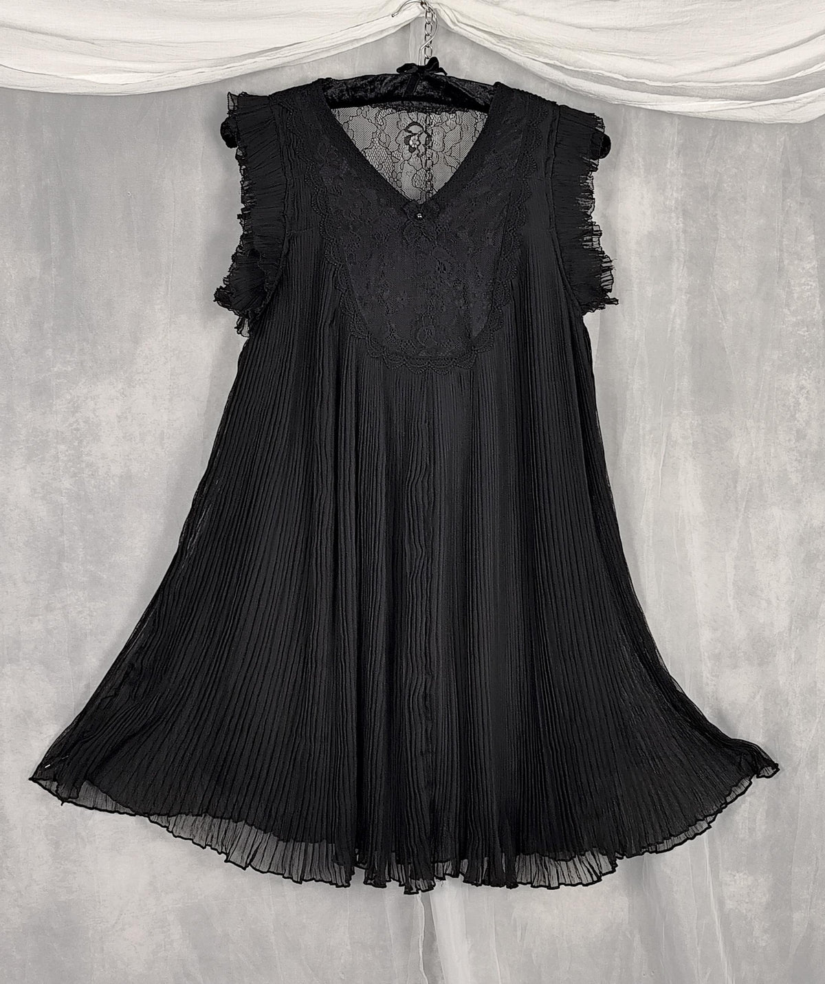 Front view of a black sleeveless pleated dress with ruffled cap sleeves and a lace V-neck bodice, hanging on a black hanger against a gray draped fabric backdrop.