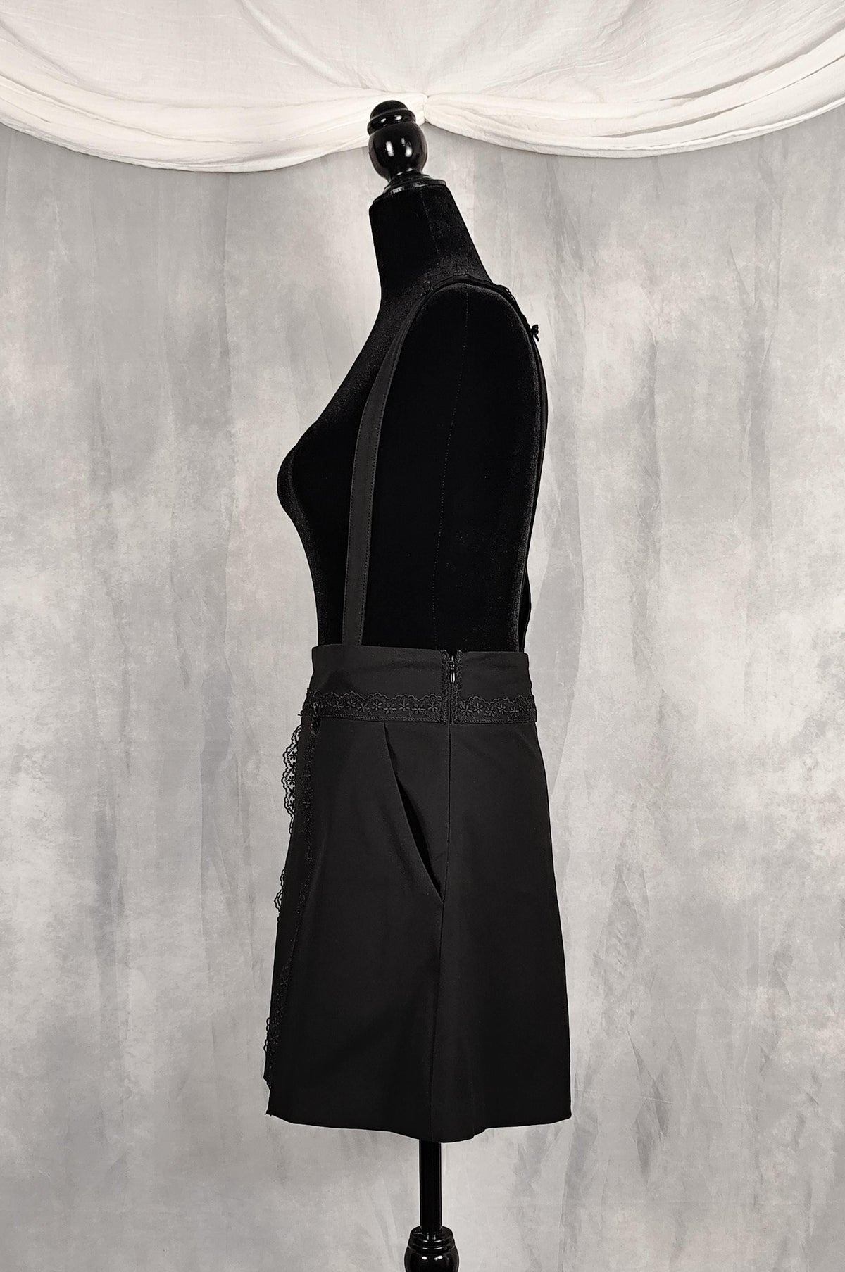 Side view of a high knee black skirt with removable straps and lace and satin bows details. It has functional pockets at front and hidden zipper.