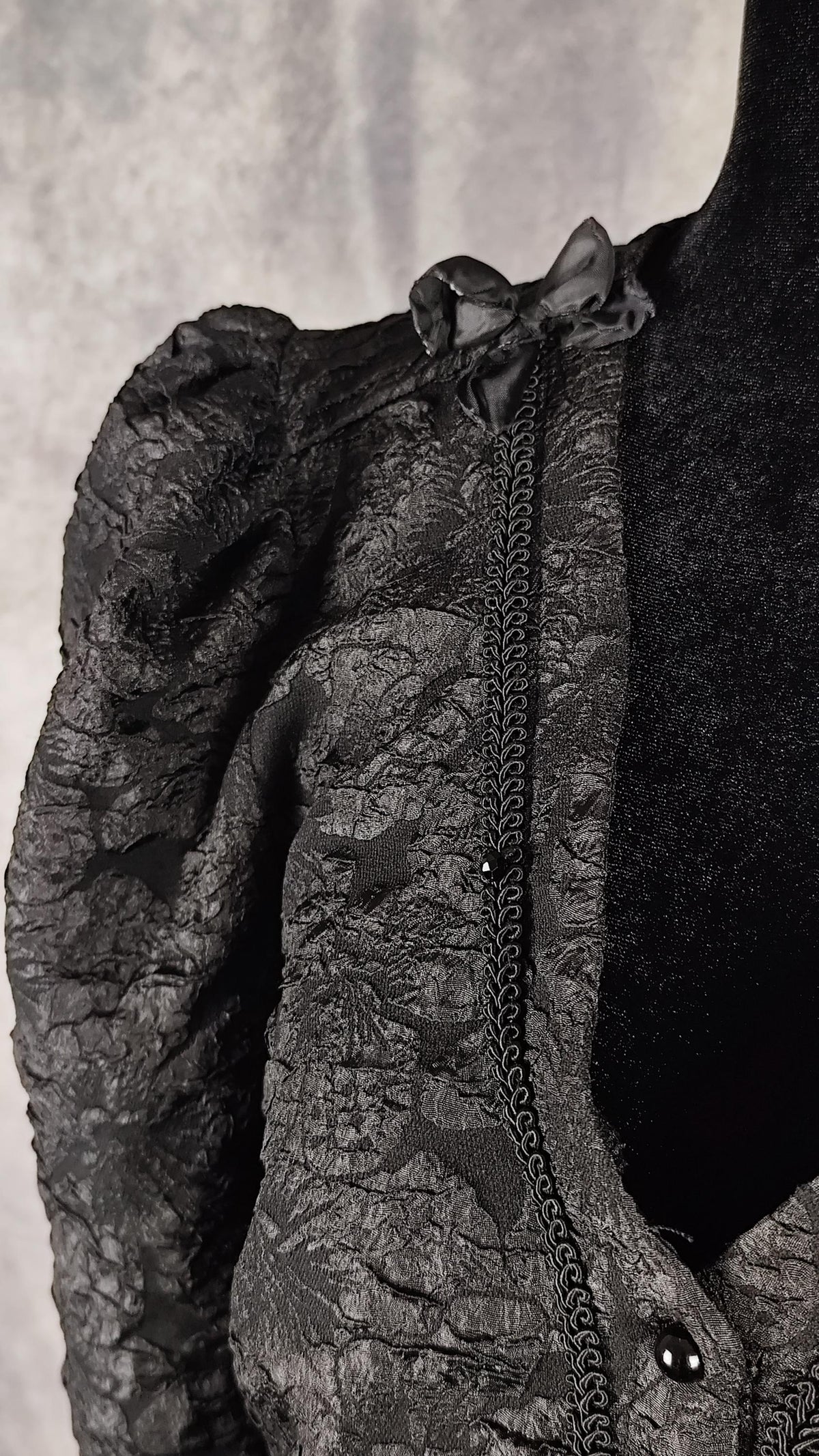 Cliose-up of a black floral brocade blouse with black pearl-shaped buttons and braid trim, faceted beads and a bow.