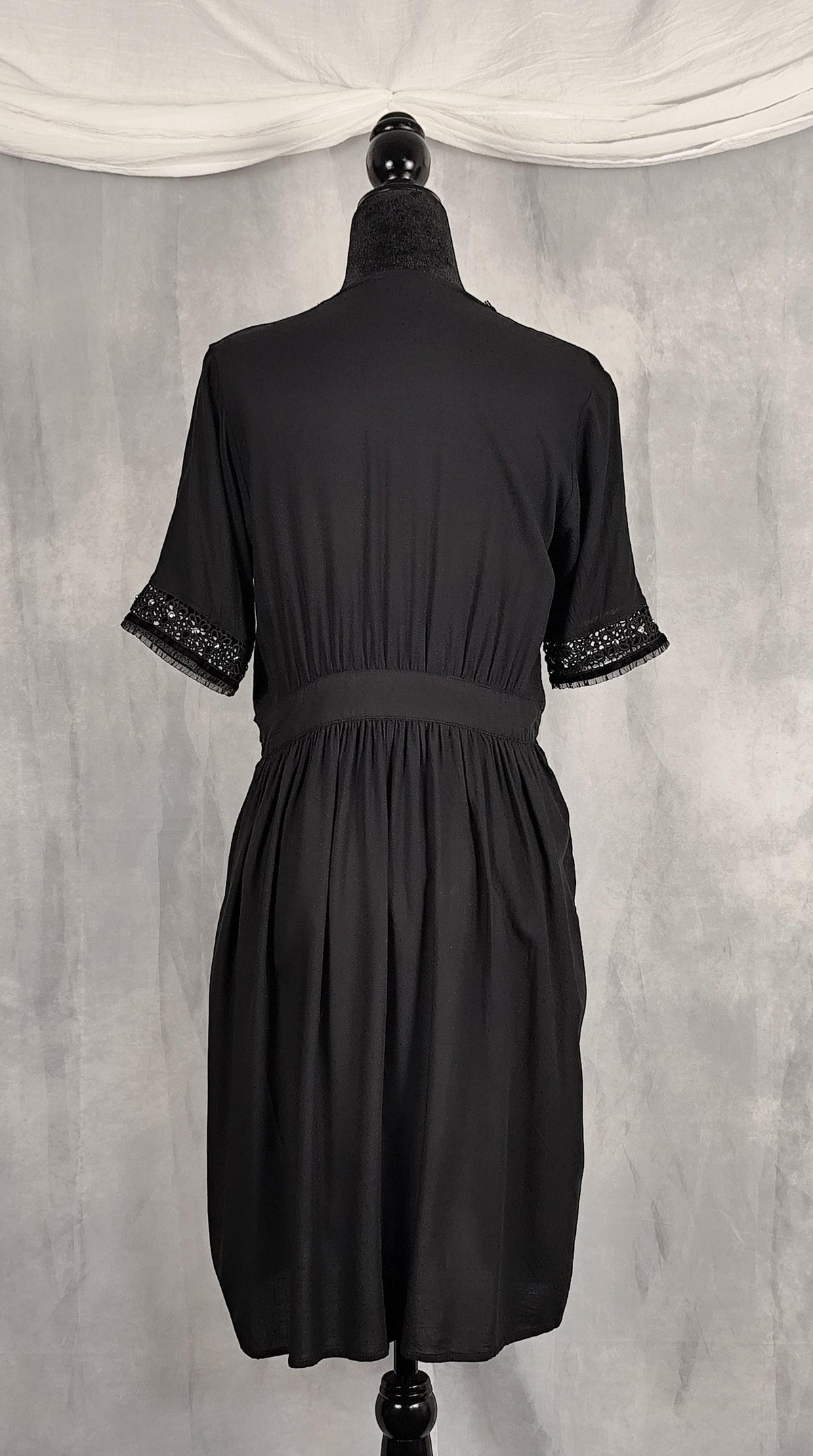 Back view of a knee high black short-sleeve wrap dress with decorative silver beaded trim along the V-neckline and sleeve cuffs, trimmed with velvet and tulle.