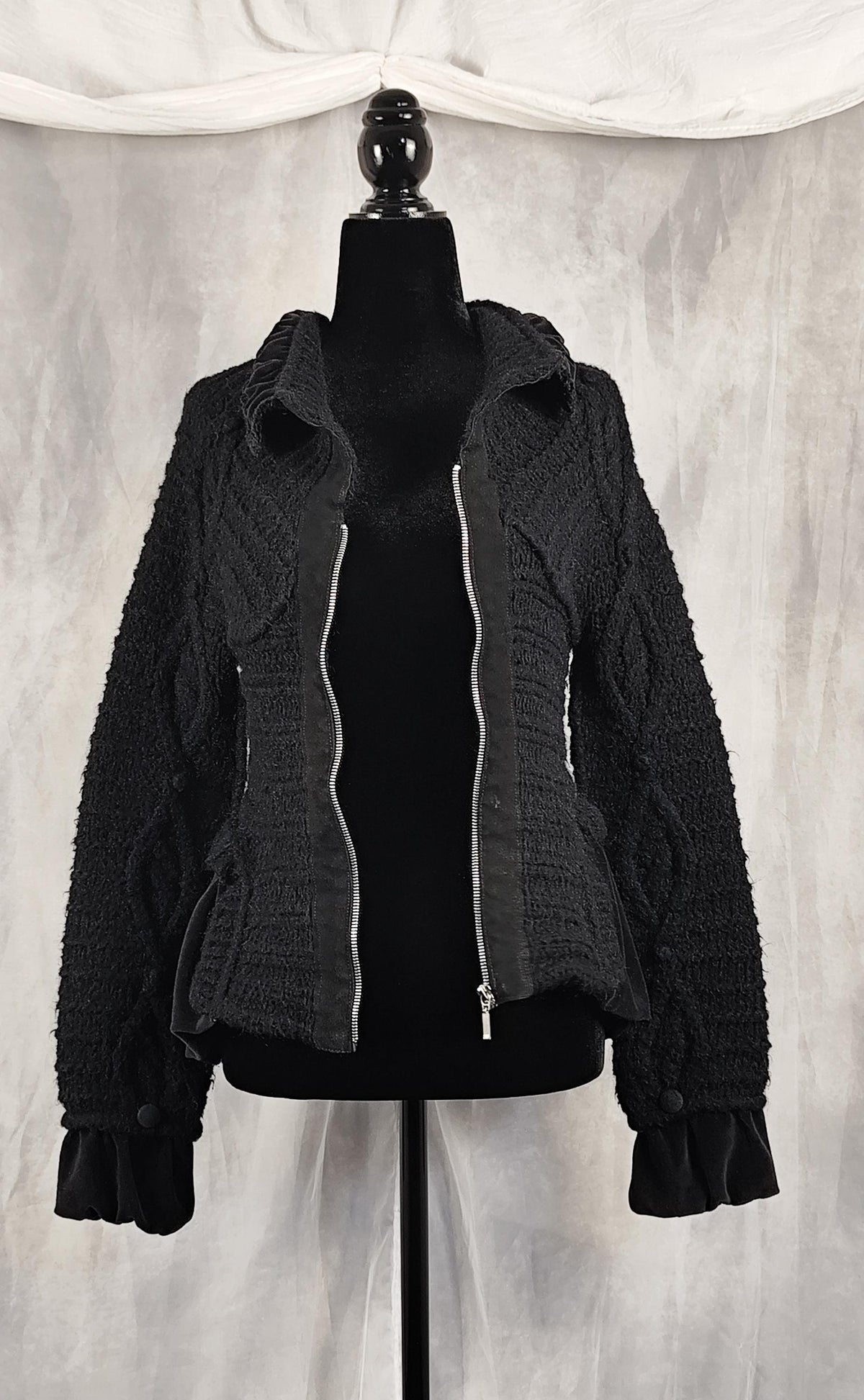 A black textured knit zip-up knit jacket with a hood and silver zipper trim, displayed on a black mannequin torso against a draped gray fabric backdrop.