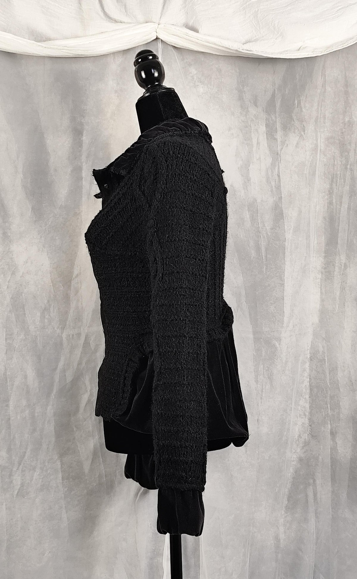 Side view of a black textured knit and velvet jacket displayed on a black dress form mannequin against a gray draped fabric backdrop.