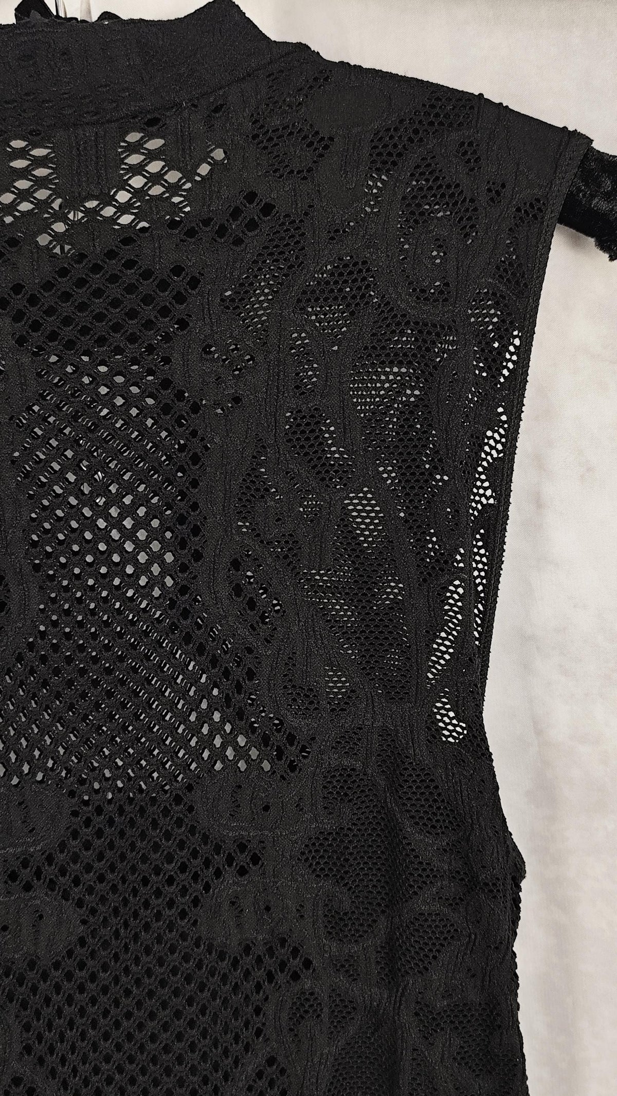 Close-up of a black sleeveless dress with a high neck and intricate floral mesh pattern, shown against a light background.