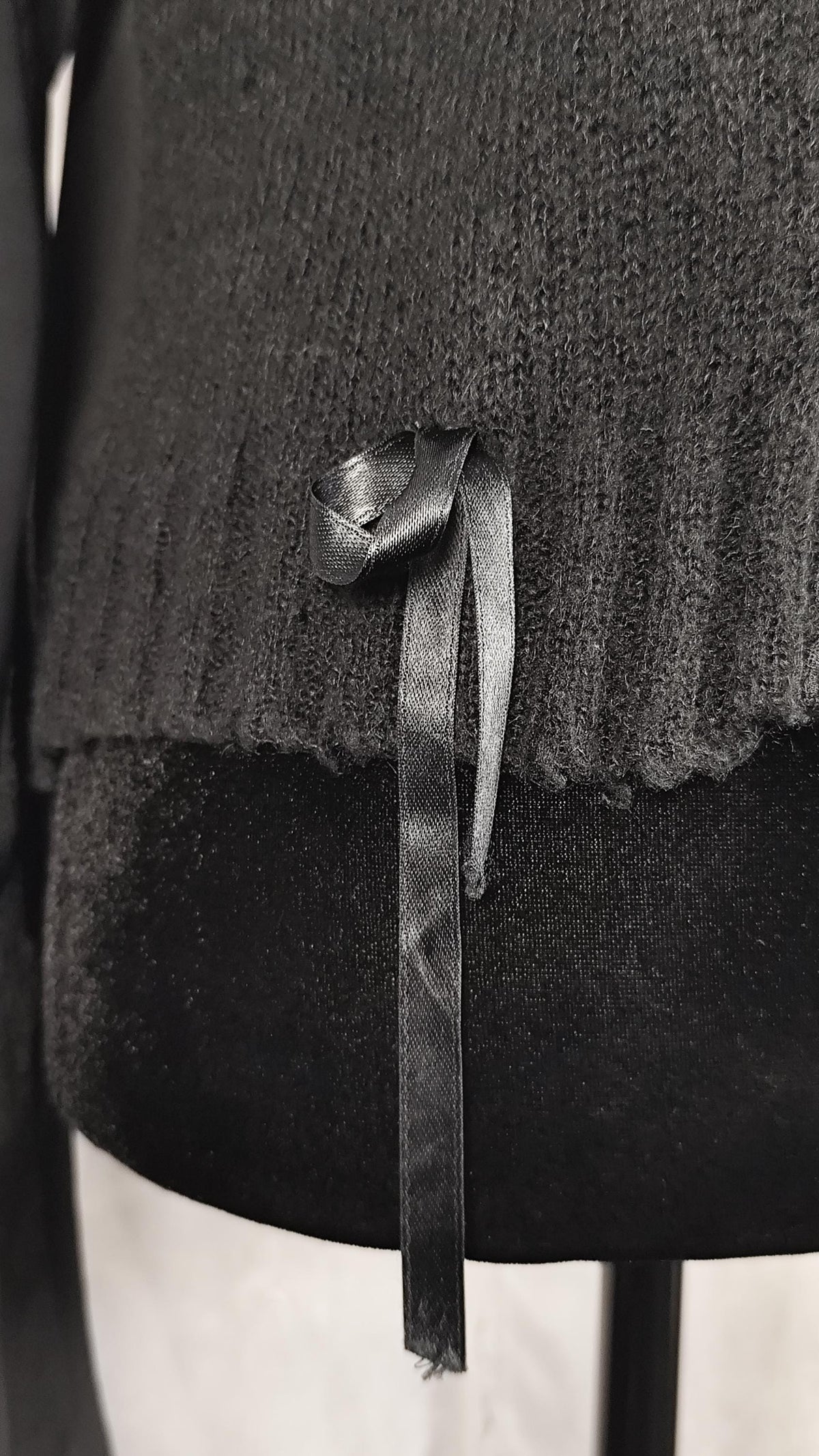 A close-up of a black ribbed knit cardigan, featuring a black satin ribbon tied in a bow and hanging from the hem.