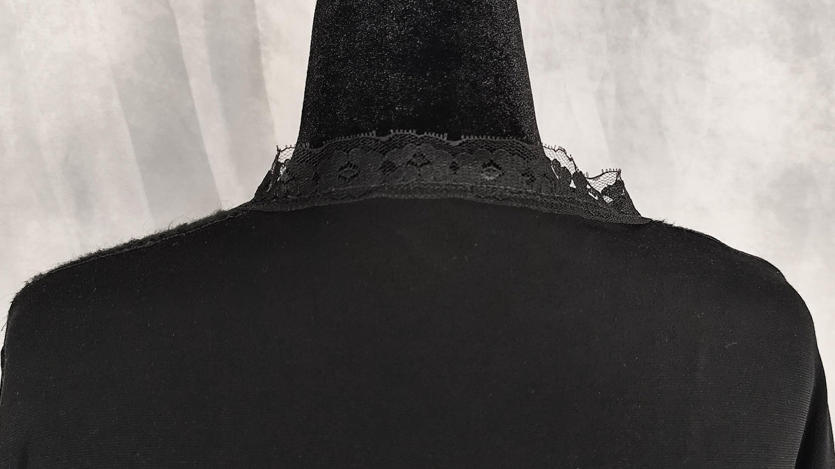 Close-up of the back of a black garment on a mannequin, showing a wide black collar with delicate lace trim against a light gray textured background.