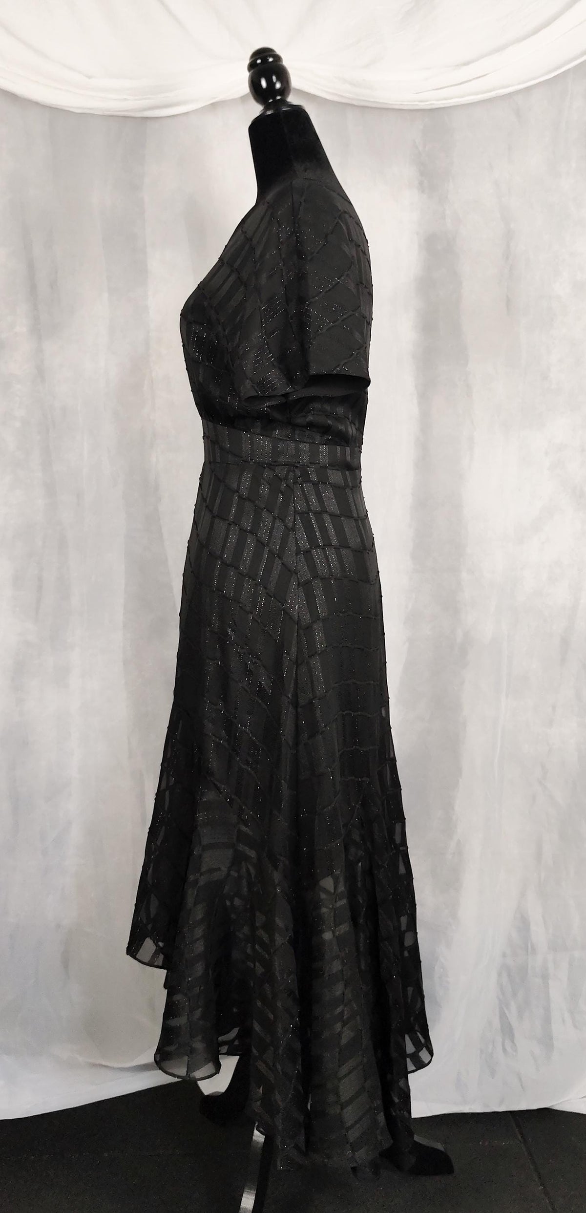 Side view of a black lace and mesh maxi dress displayed on a black mannequin against a draped gray fabric backdrop.