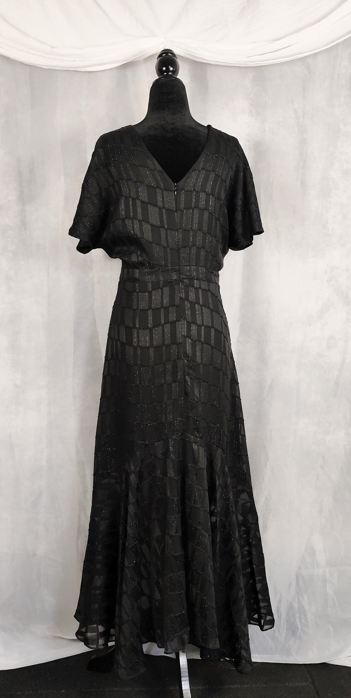 Back view of a black short-sleeved maxi dress with a textured geometric pattern, displayed on a black mannequin against a draped white fabric backdrop.