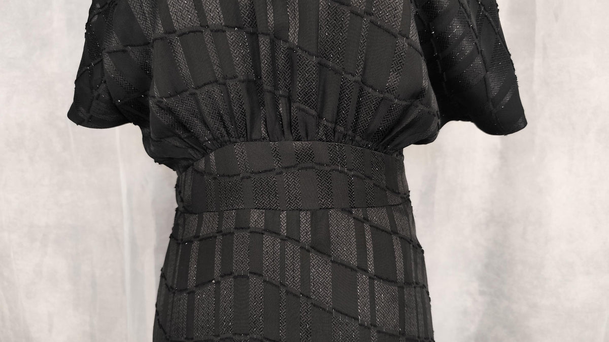 A close-up of a black textured dress with a geometric pattern and a matching waistband, shown on a mannequin against a neutral gray background.