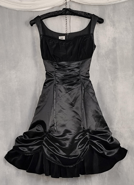 Front view of a black satin and velvet dress with a corset-style bodice and ruffled skirt, hanging on a black hanger against a textured gray backdrop.