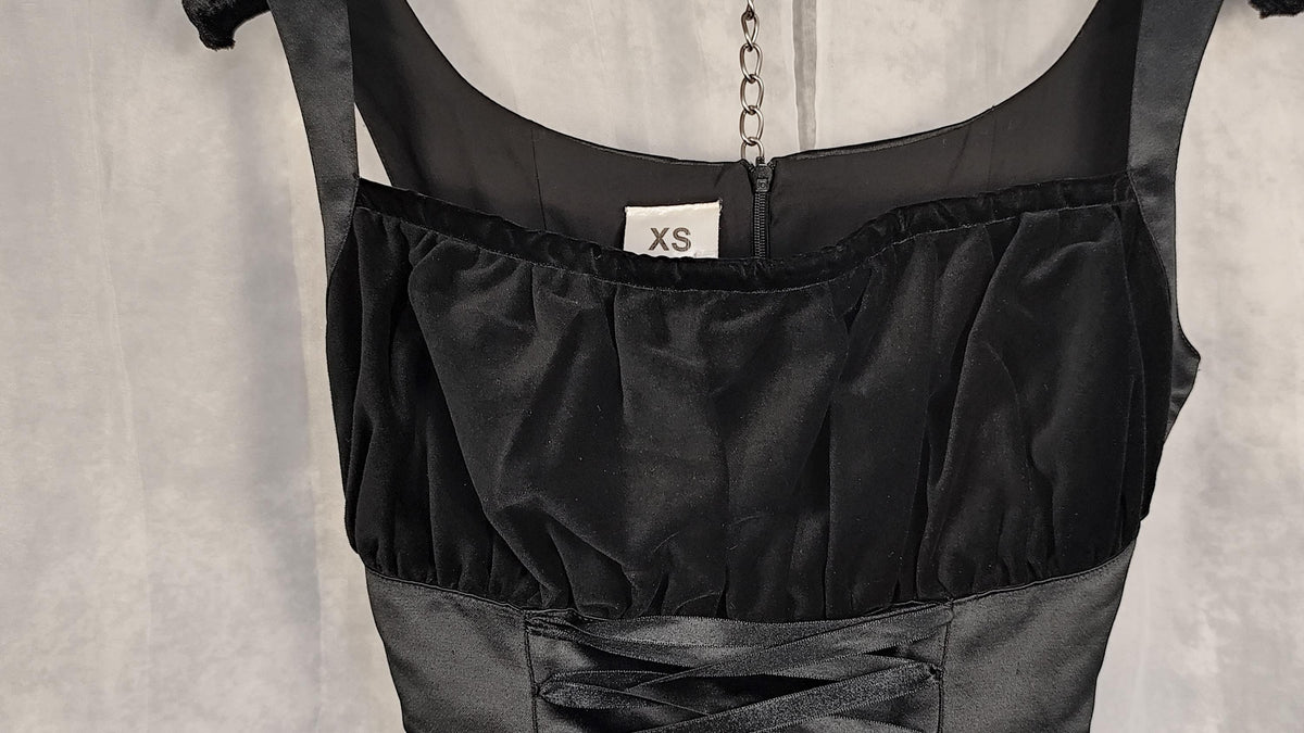 Close-up of a black satin-like garment with velvet ruched detailing and a small white size label reading XS, shown against a light gray fabric background.
