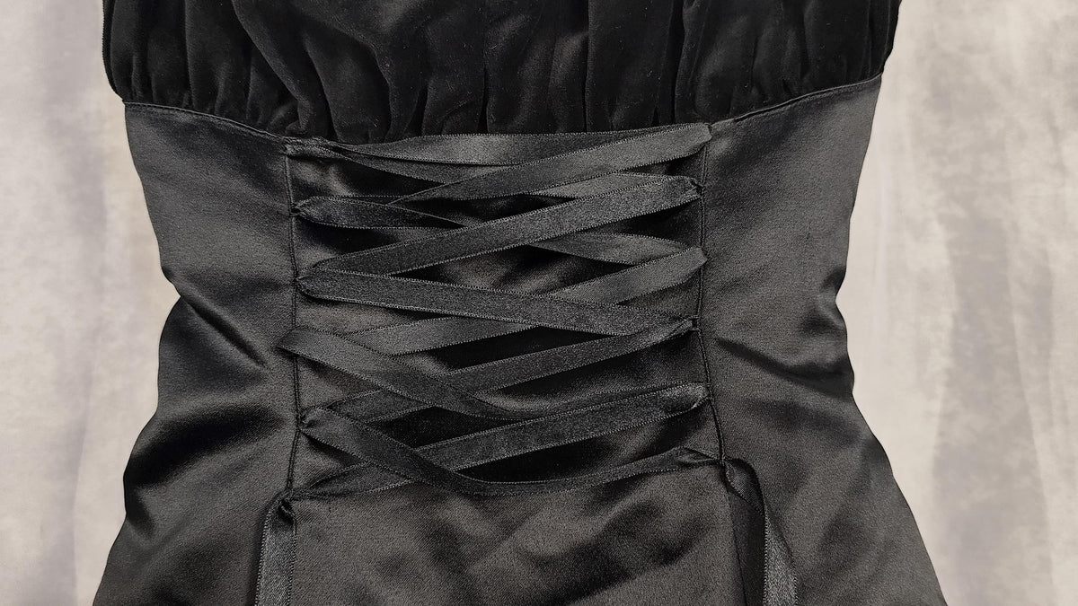 Close-up of a black satin corset-style dress waist with crisscross lace-up straps against a textured gray background.