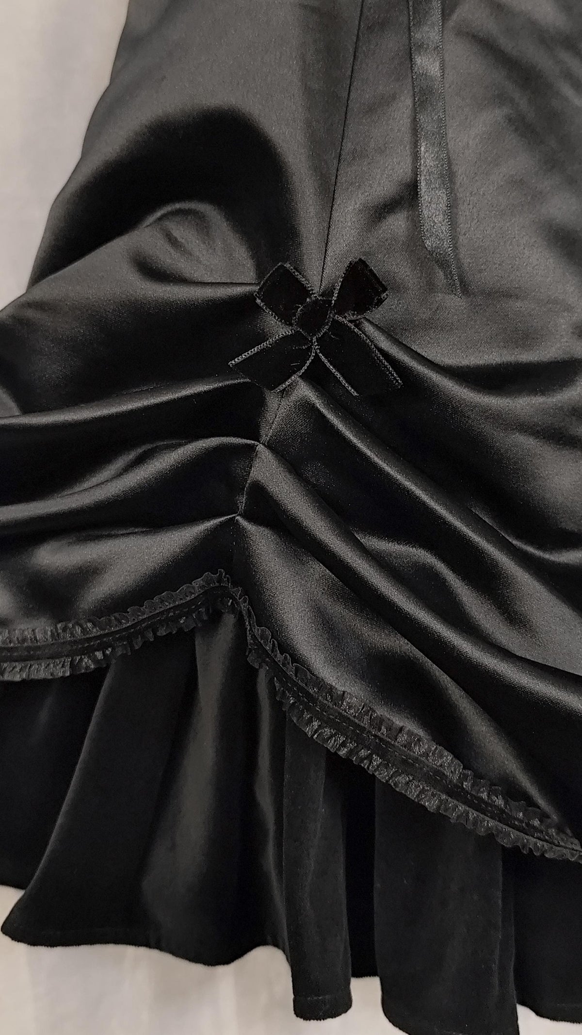 Close-up of a black satin dress with gathered draping, a small vekvet bow detail at the center, and a ruffled hem against a light neutral background.