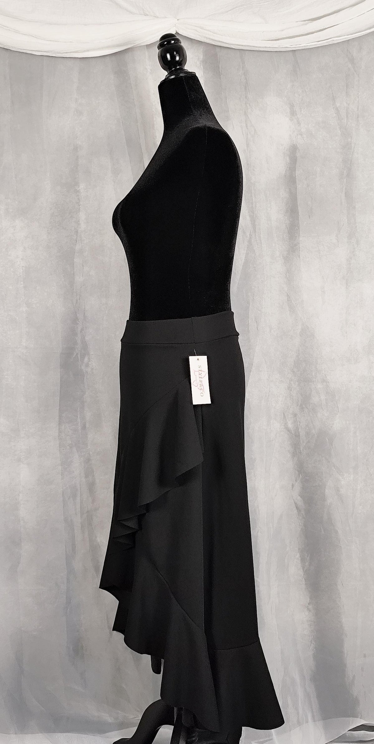 Side view of a black ruffled asymmetrical skirt displayed on a black dress form mannequin against a gray draped fabric backdrop.