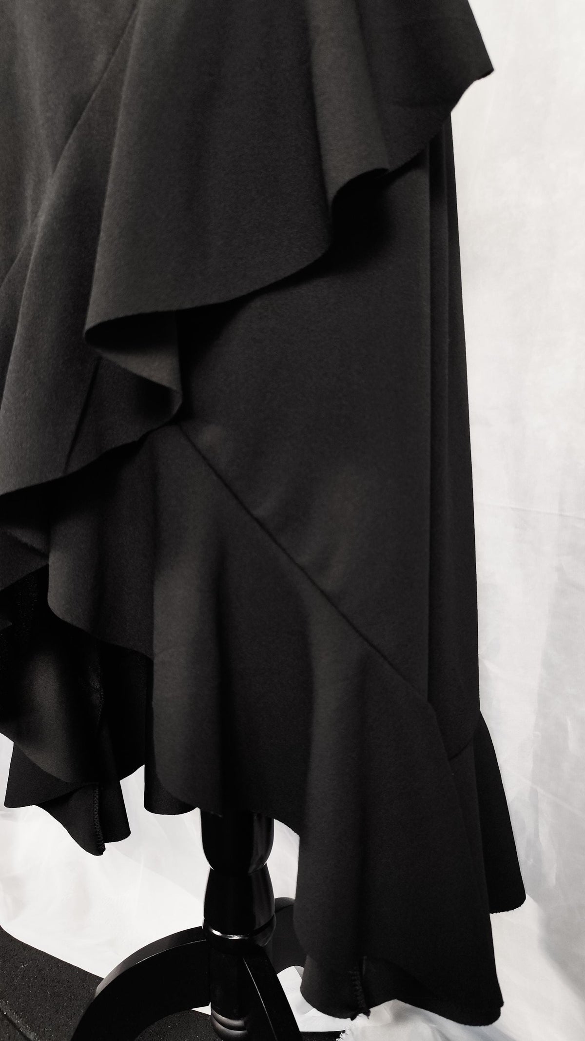 A close-up of a black ruffled skirt displayed on a mannequin stand against a white studio backdrop.