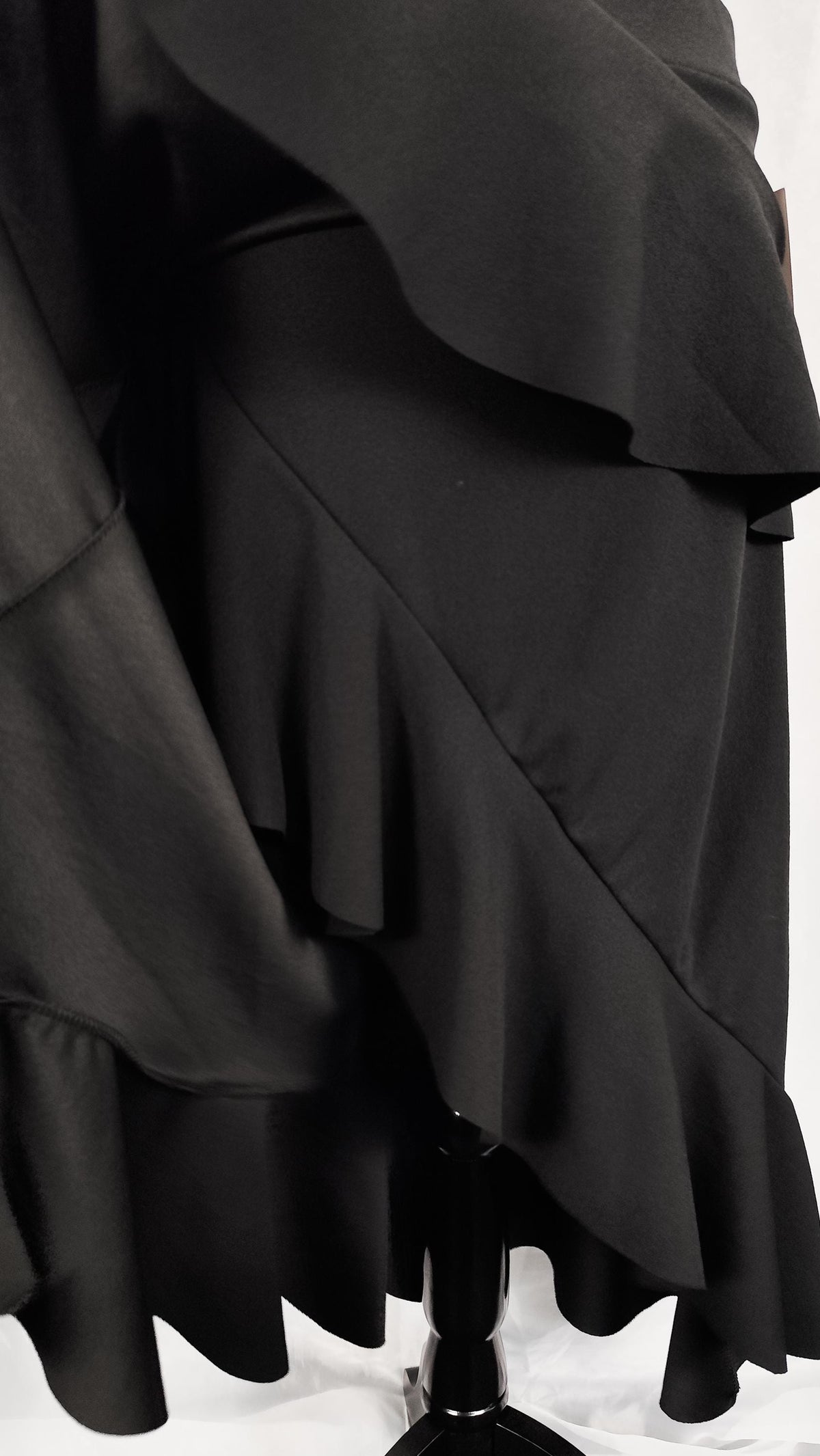 A close-up of a black ruffled skirt displayed on a black mannequin stand against a white studio background.