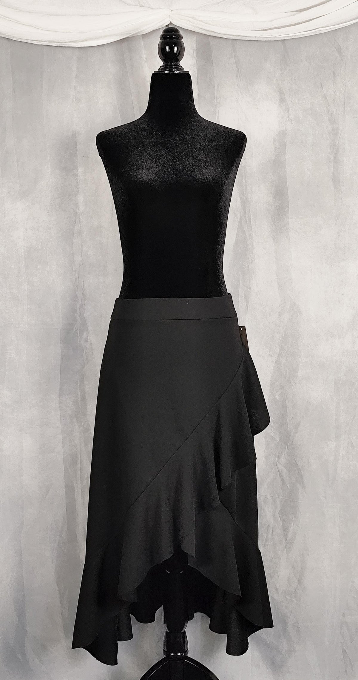 Front view of a black ruffled wrap-style skirt displayed on a black mannequin torso against a textured gray backdrop with draped white fabric at the top.
