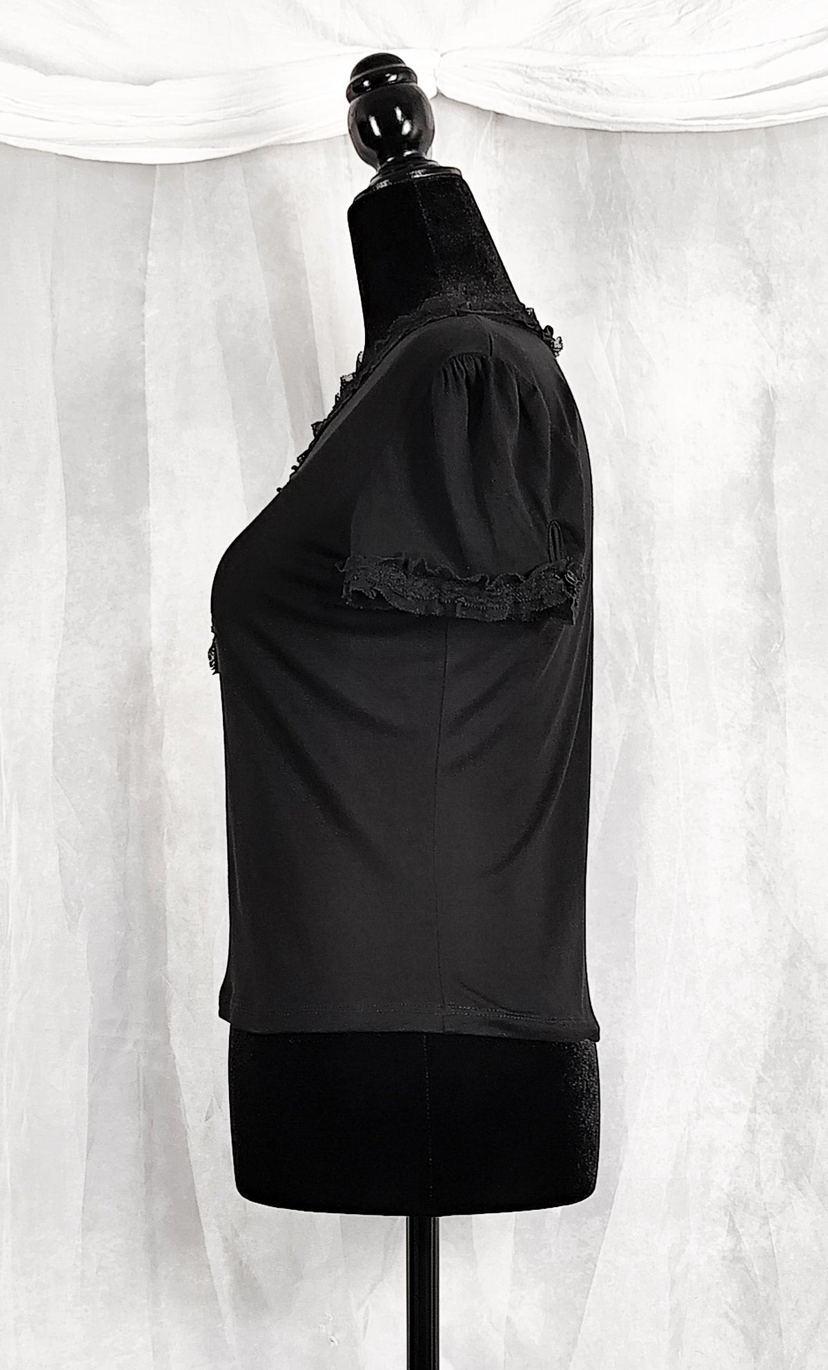 Side view of a black short-sleeve blouse with ruffled trim on the shoulders and upper chest, displayed on a black dress form against a white draped fabric backdrop.