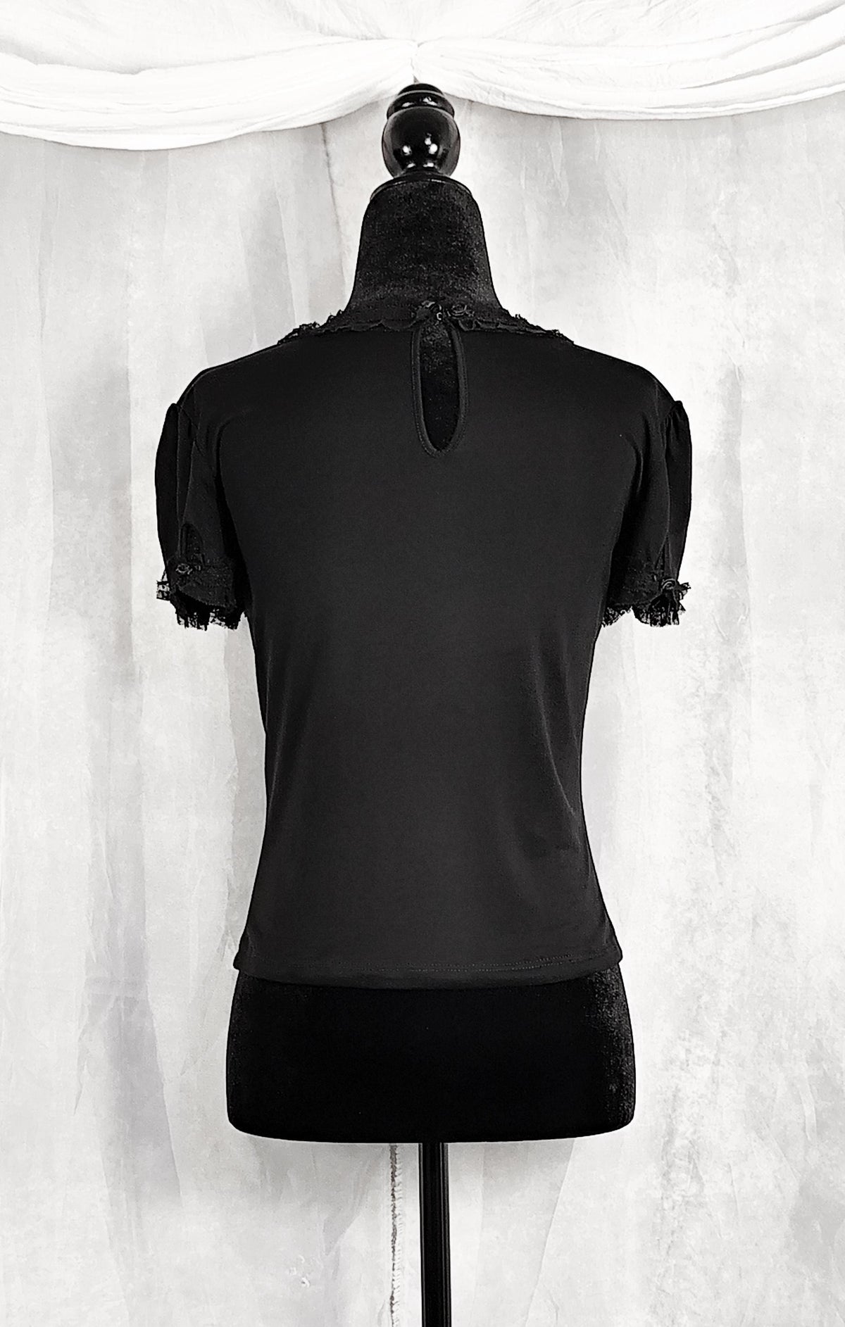 Back view of a black short-sleeve top displayed on a black dress form mannequin, showing the back view with a small keyhole cutout and lace trim at the neckline and sleeve hems, set against a white draped fabric backdrop.