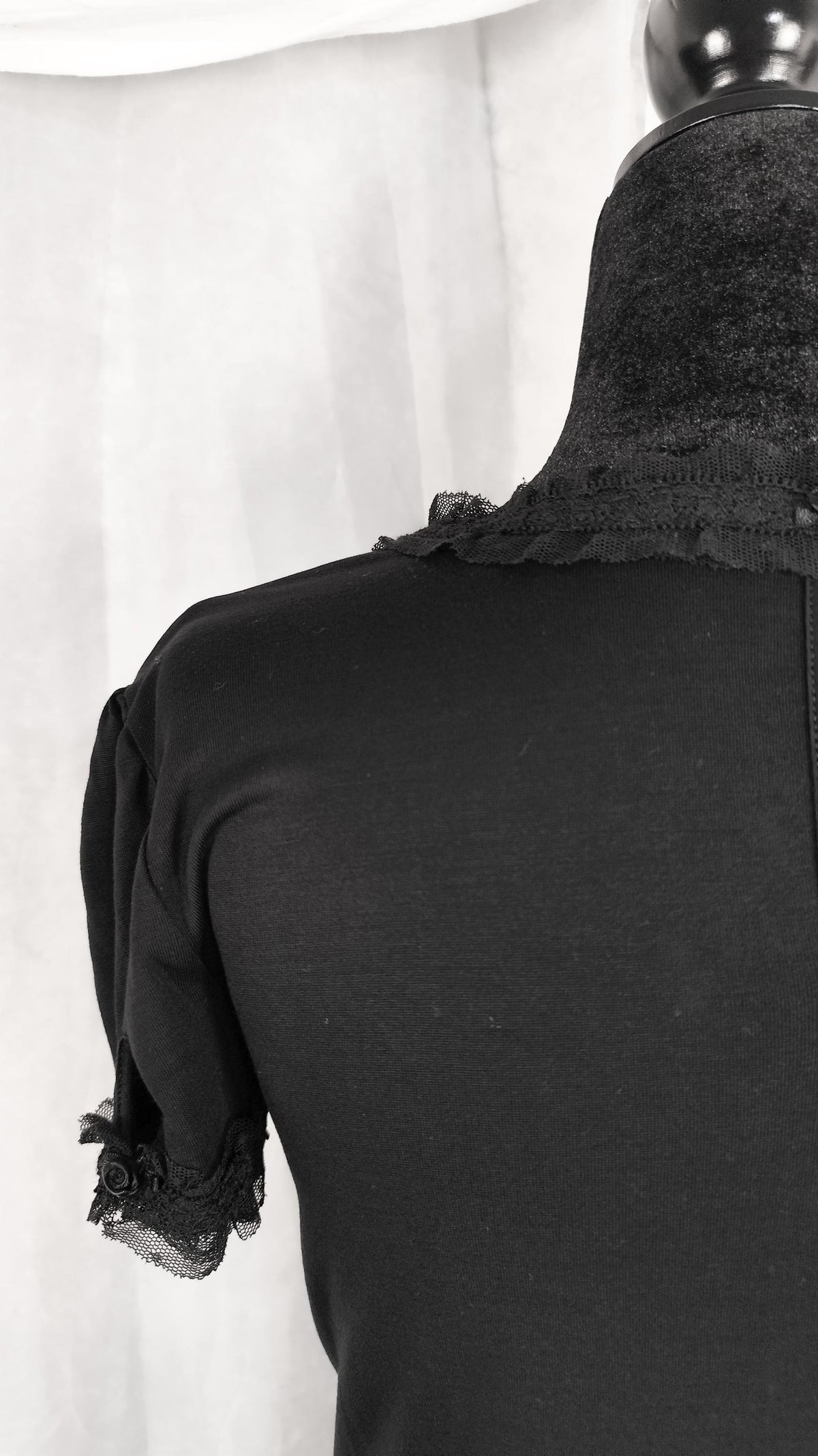 A black short-sleeve top displayed on a mannequin from the back, featuring ruffled lace trim at the neckline and sleeve cuffs, set against a light draped fabric backdrop.