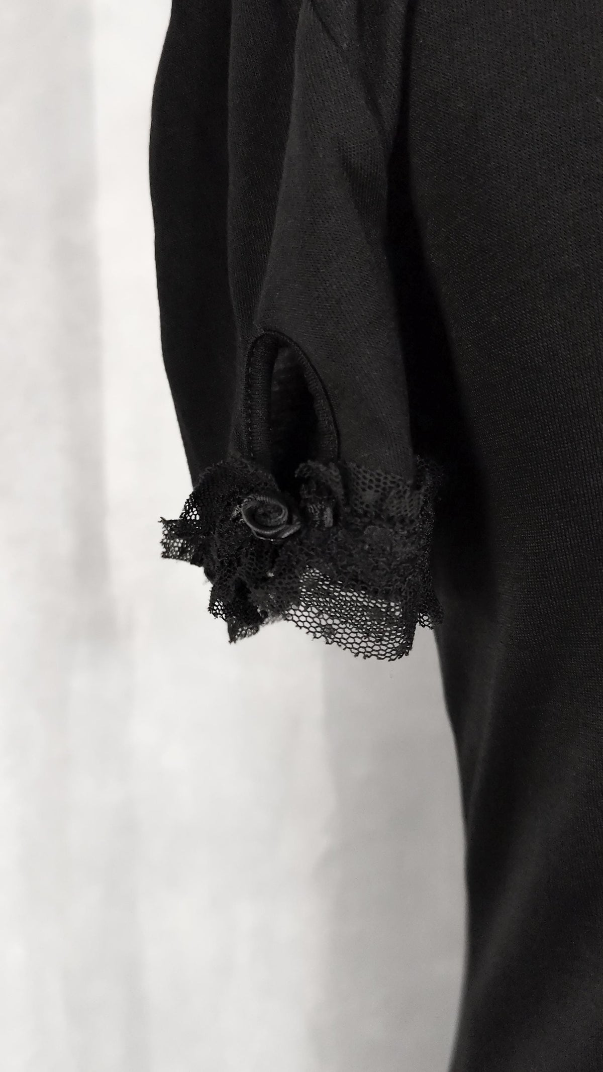 Close-up of a black top sleeve featuring a ruffled lace cuff with a small black fabric rose detail, set against a light gray background.