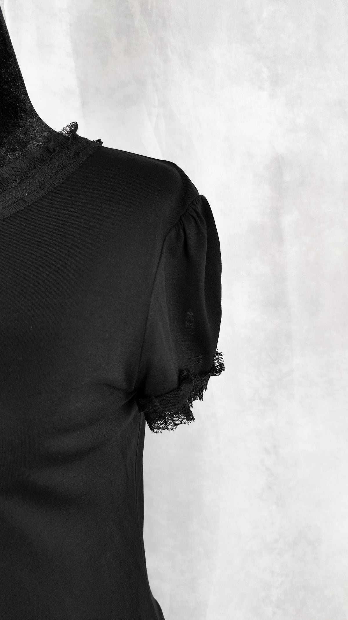 A close-up of a black short-sleeve blouse on a mannequin, featuring sheer black lace trim at the collar and sleeve cuffs, set against a textured light gray background.