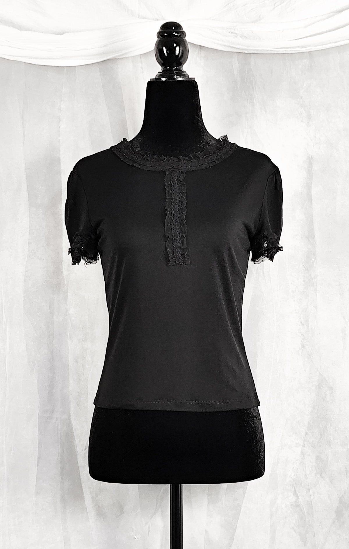 Front view of a black short-sleeve top with lace trim at the collar and sleeve cuffs, displayed on a black dress form mannequin against a white draped fabric backdrop.