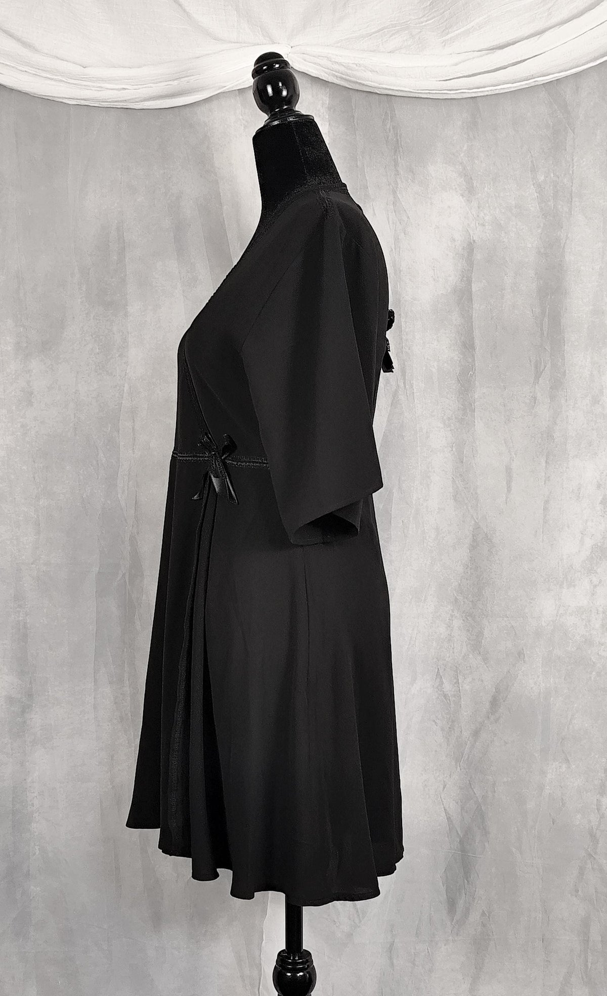 Side view of a black short-sleeve dress displayed on a black mannequin against a textured gray backdrop with draped white fabric at the top.