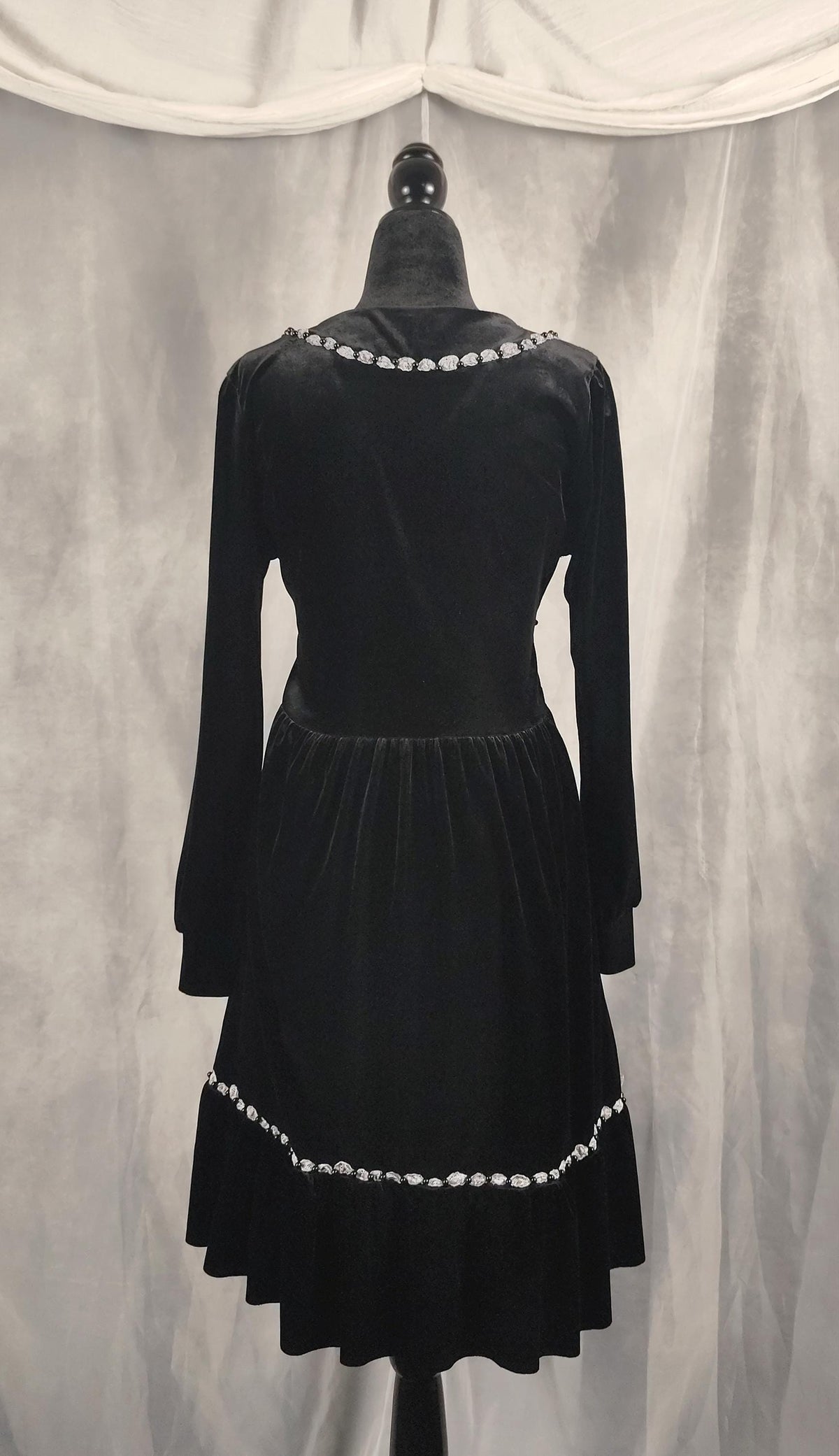 Back view of a black velvet long-sleeve dress displayed on a black dress form against a gray draped fabric backdrop. The dress features a flared skirt and is decorated with a row of white pearls around the neckline and a matching pearl-trimmed hem.
