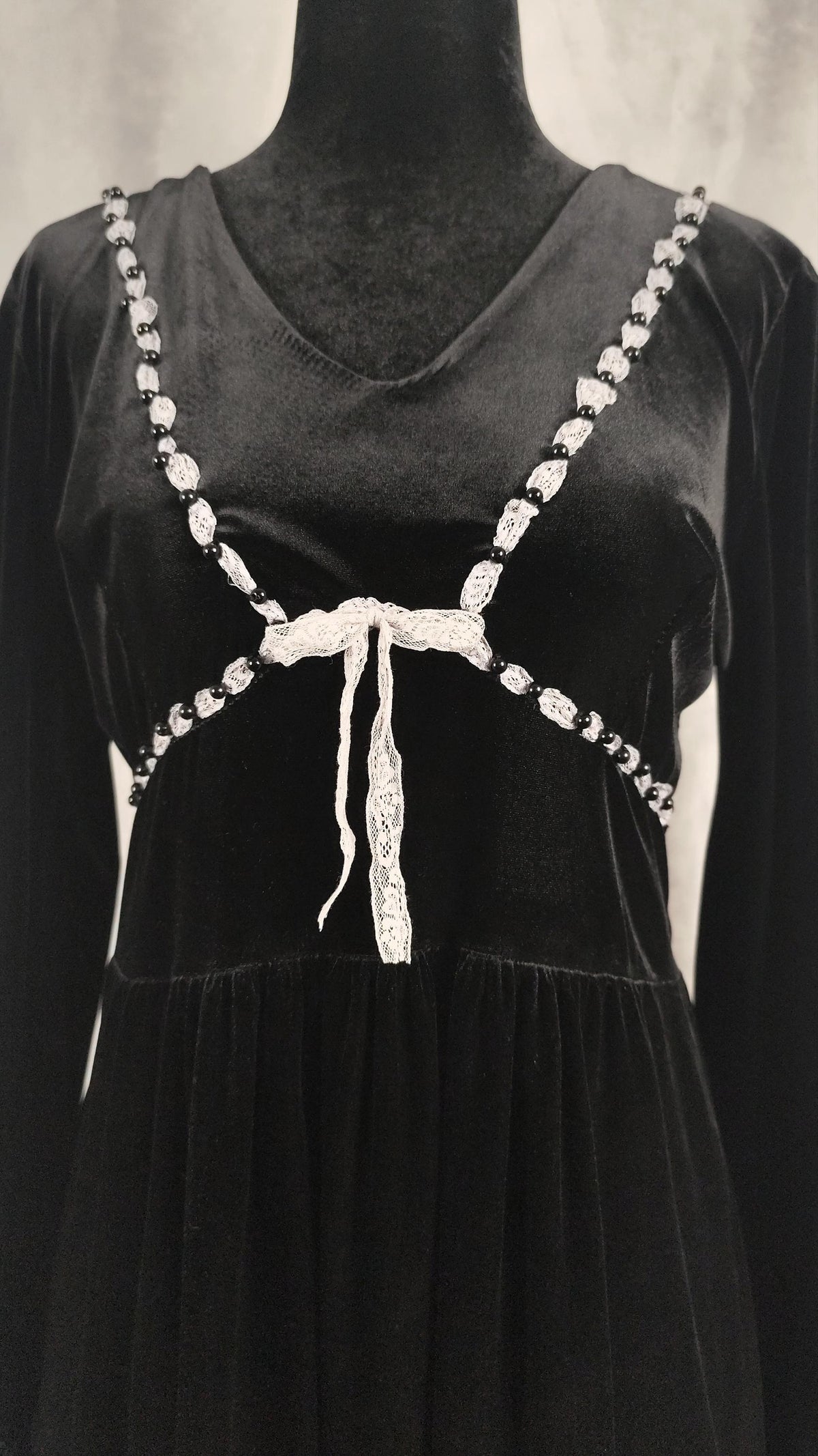 Close-up of a black velvet long-sleeve dress displayed on a black mannequin torso, featuring an off-white beaded halter-style strap design with a central bow and dangling tie details.